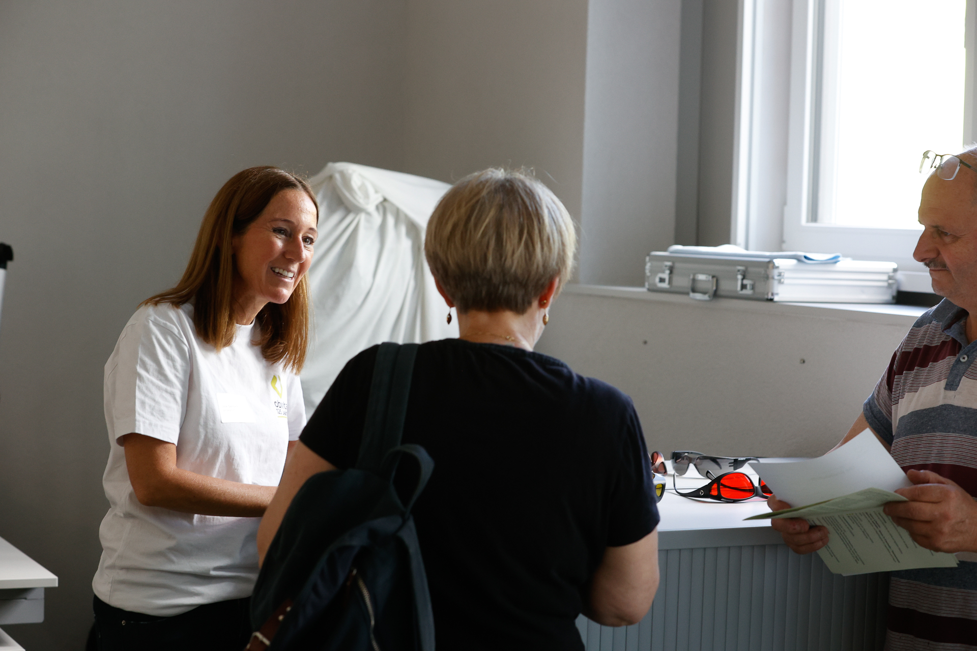 Eine Frau in einem weißen Hemd lächelt, während sie zwei Personen an einem Schalter in einem hell beleuchteten Büro bedient; auf dem Schalter liegen Unterlagen und eine Brille.