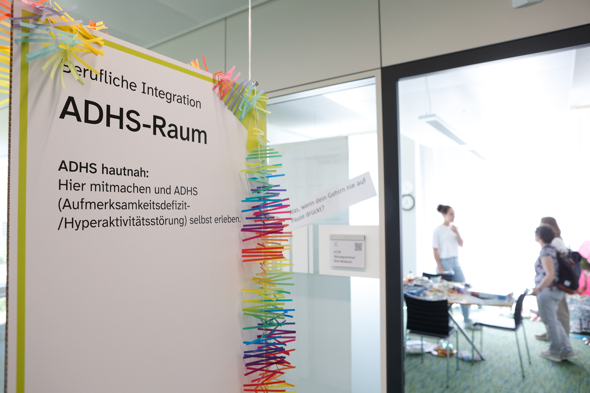Am Eingang eines glaswandigen Raums steht ein Schild mit der Aufschrift „ADHS-Raum“ und bunten Wäscheklammern. In diesem hellen, modernen Büro unterhalten sich mehrere Personen an einem Tisch.