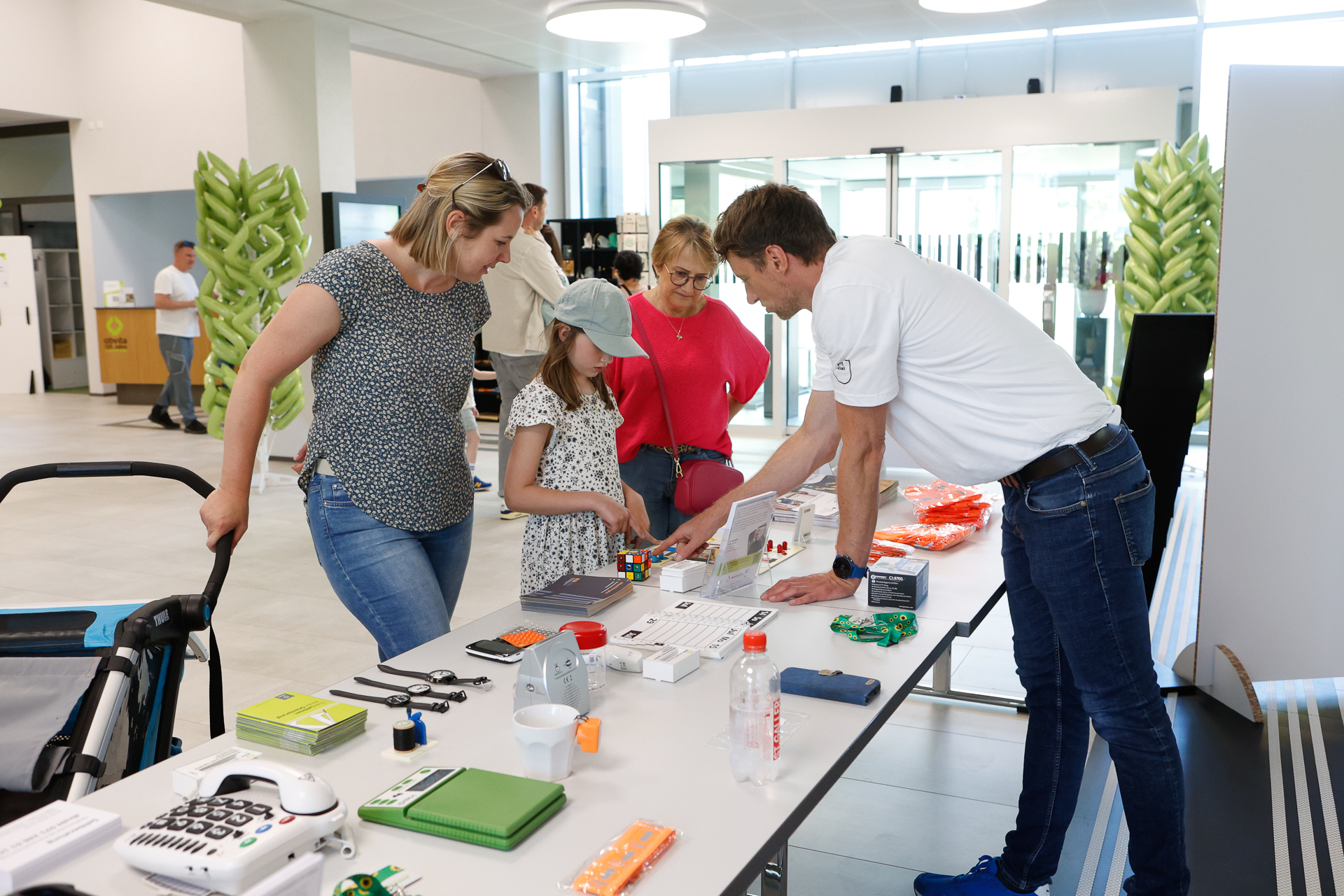 In einem hellen, modernen Innenraum erklärt ein Mann drei Personen, darunter ein kleines Mädchen, verschiedene Gegenstände auf einem Ausstellungs-Tisch. Auf dem Tisch liegen technische Geräte, Werkzeuge und Informationsmaterialien. Am Ende des Tisches ist ein Kinderwagen zu sehen.