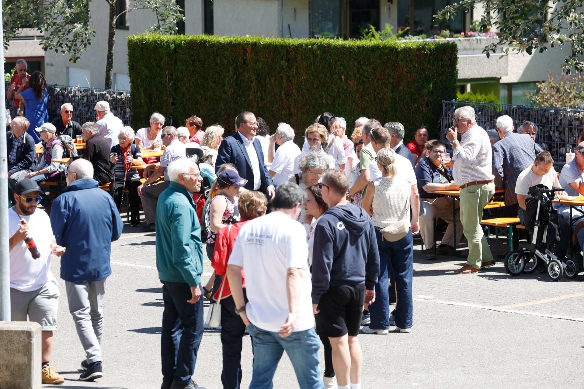 An einem sonnigen Tag versammelt sich eine große Gruppe von Menschen, hauptsächlich Erwachsene und Senioren, im Freien; sie sitzen an orangefarbenen Tischen oder stehen im Gespräch beieinander – offenbar handelt es sich um eine Gemeinschaftsveranstaltung oder ein geselliges Beisammensein.