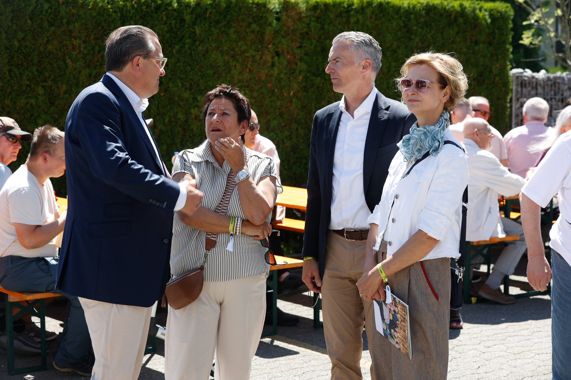 Vier Erwachsene stehen an einem sonnigen Tag im Freien und unterhalten sich; sie sind halbformell gekleidet. Im Hintergrund sind Picknicktische mit sitzenden Personen zu sehen, und grüne Hecken säumen den Bereich.