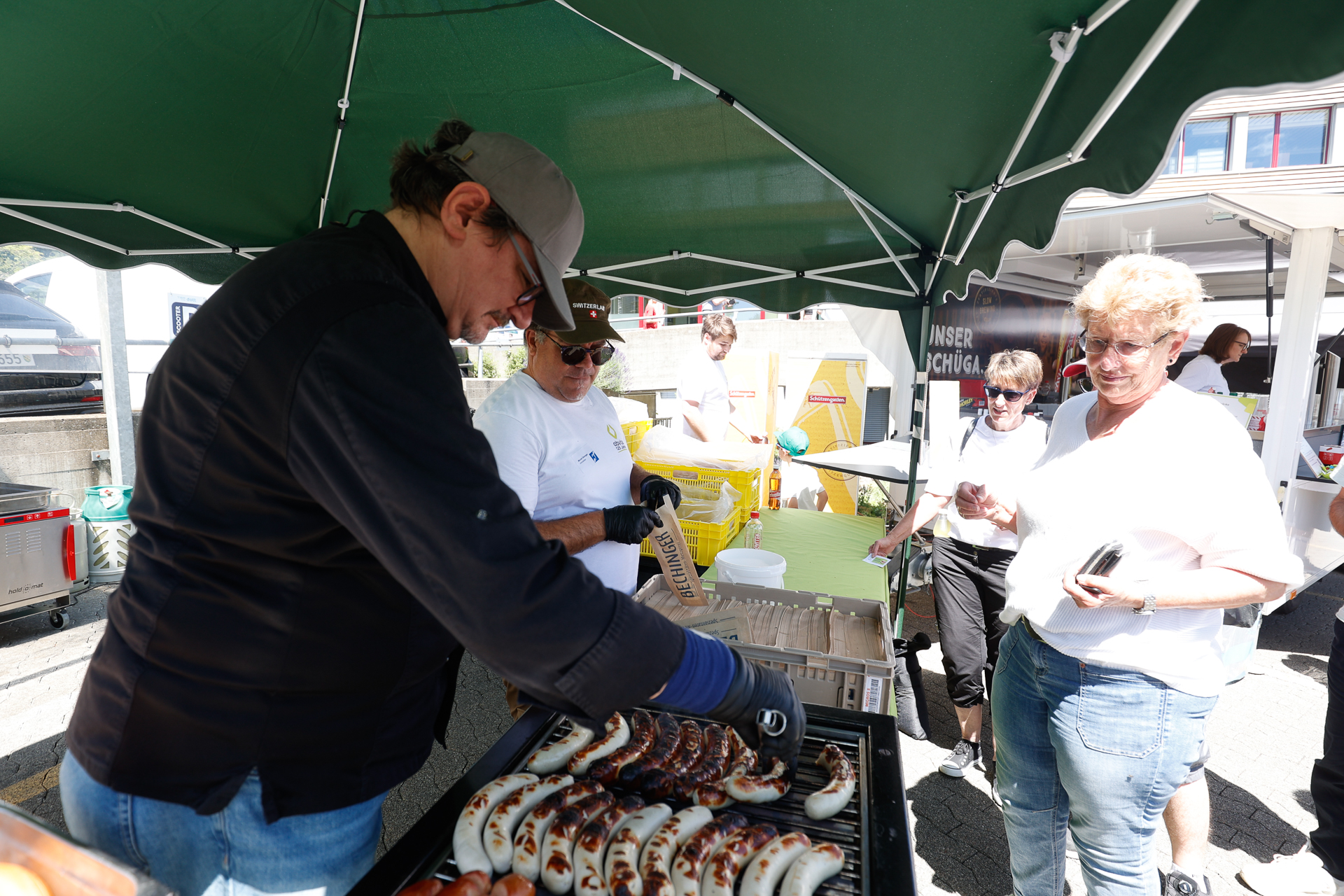 Eine Person grillt auf einem Markt unter freiem Himmel unter einem grünen Vordach Würstchen, während andere in der Nähe stehen – einige lächeln und warten darauf, bedient zu werden. Im Hintergrund sind Tische und Marktwaren zu sehen.