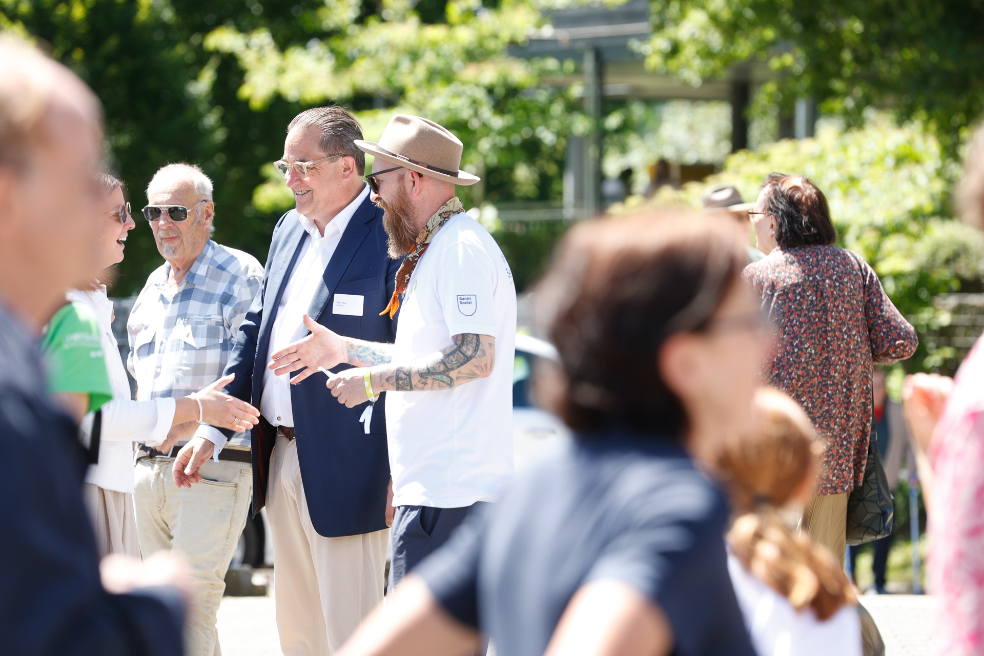 Eine Gruppe von Menschen, die sich an einem sonnigen Tag im Freien aufhalten; einige unterhalten sich. Im Hintergrund sind Bäume und ein Gebäude zu sehen. Die Atmosphäre wirkt ungezwungen und lebhaft.