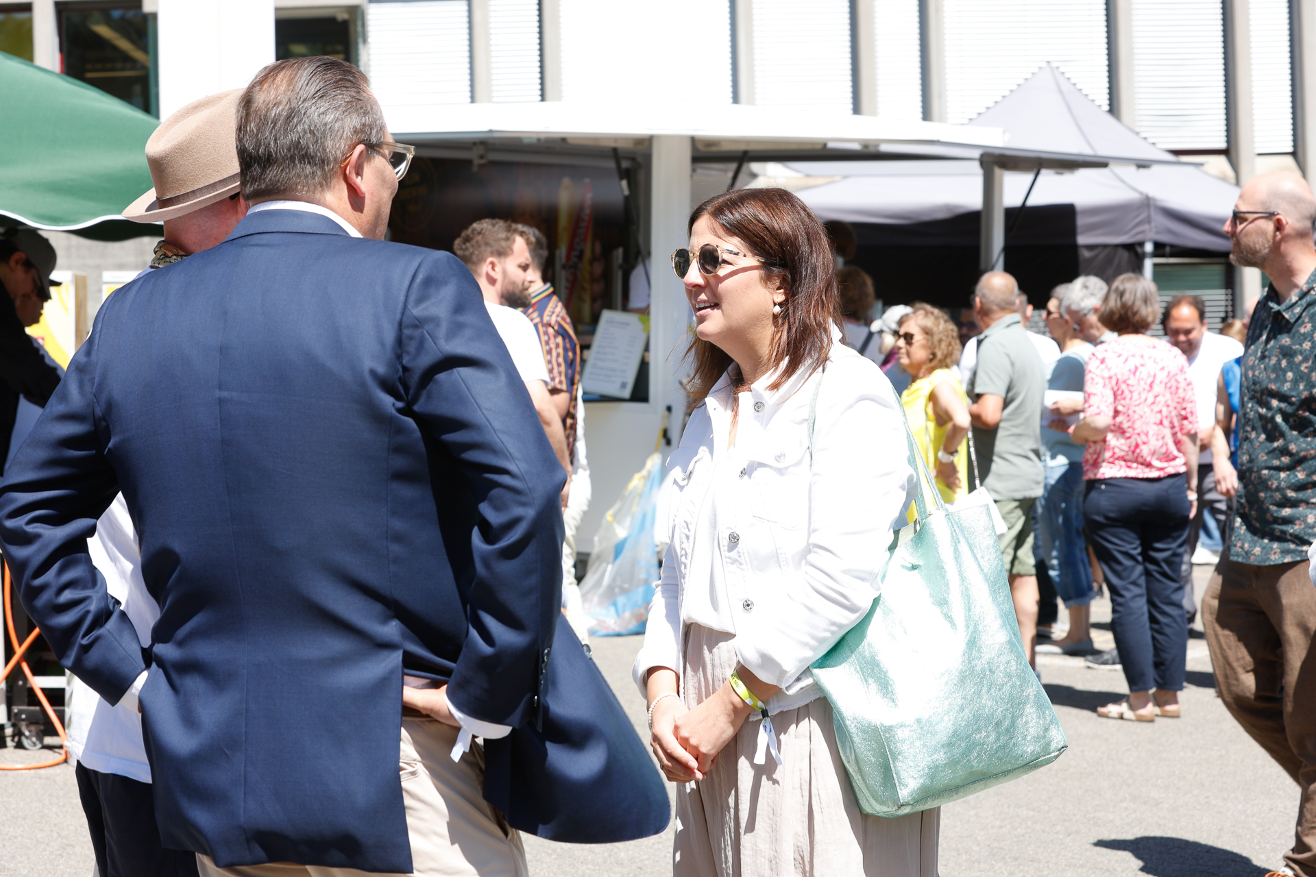 Eine Gruppe von Menschen unterhält sich im Freien bei einer sonnigen Veranstaltung oder auf einem Markt, im Hintergrund sind Stände und andere Besucher zu sehen. Eine Frau mit Sonnenbrille und weißer Jacke lächelt, während sie sich mit zwei Männern in Anzügen unterhält.