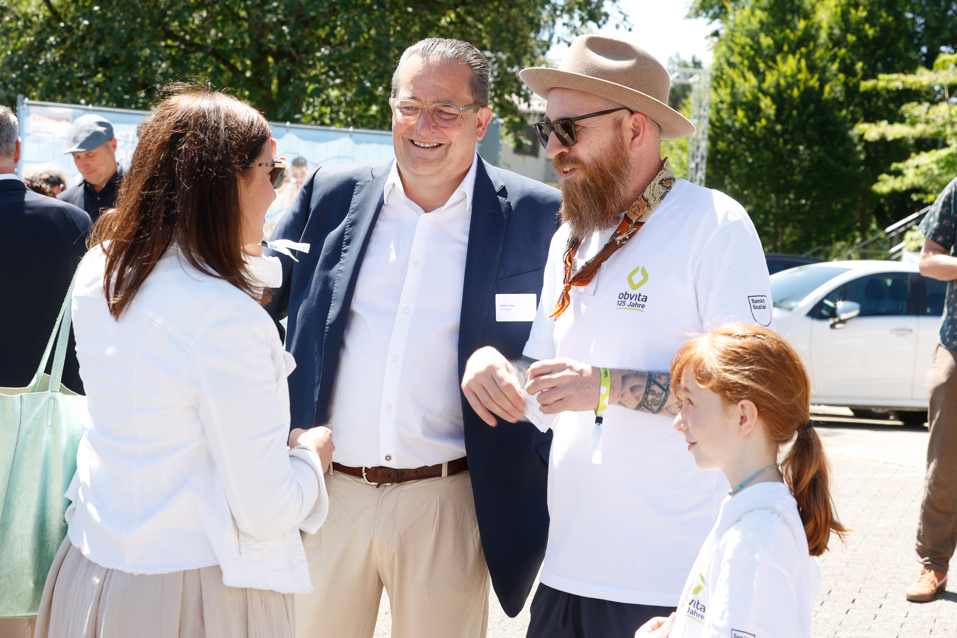 Eine Gruppe von Erwachsenen und ein Kind stehen gemeinsam im Freien, lächeln und unterhalten sich. Die Erwachsenen tragen Business- oder Freizeitkleidung, und das Kind hat rote Haare und trägt ein weißes Hemd. Im Hintergrund sind Bäume und geparkte Autos zu sehen.