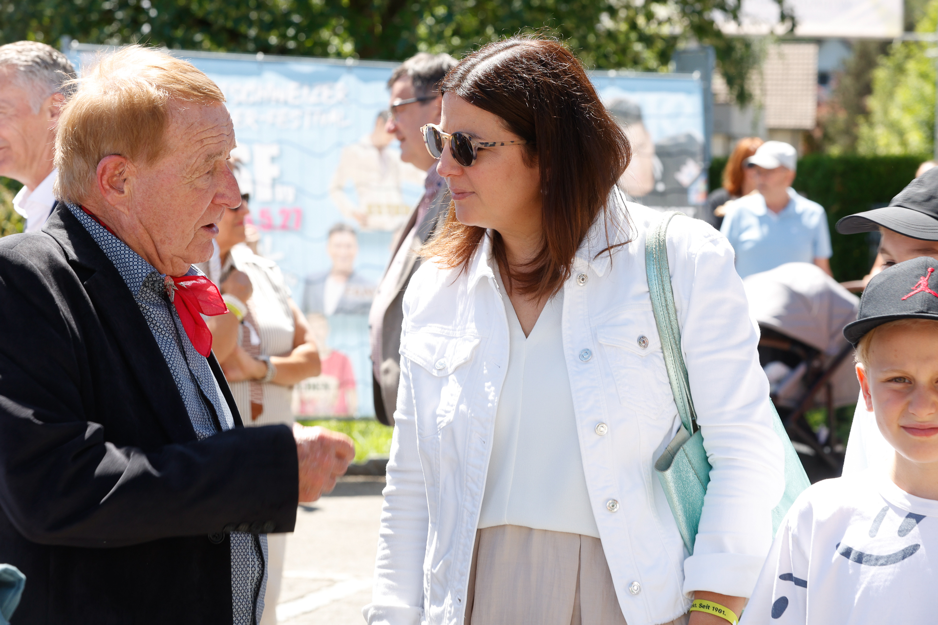 Ein älterer Mann in einer schwarzen Jacke und einem roten Schal unterhält sich bei einer Veranstaltung im Freien mit einer Frau, die eine Sonnenbrille und eine weiße Jacke trägt. Im Hintergrund sind an einem sonnigen Tag weitere Personen und ein großes Plakat zu sehen.