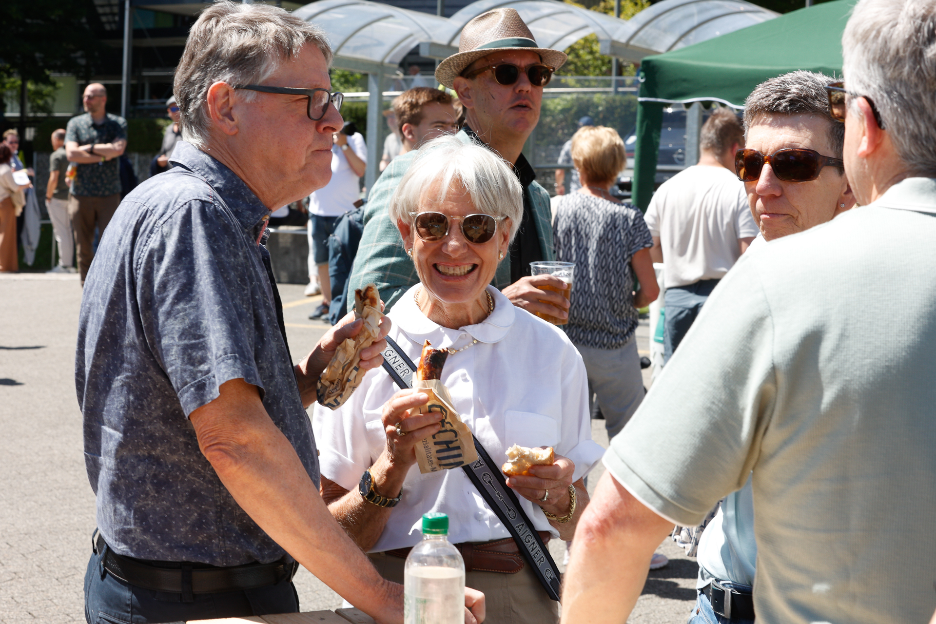 Eine Gruppe älterer Menschen steht bei einer Veranstaltung im Freien in der Sonne, lächelt und isst Würstchen im Brötchen. Eine Frau mit Sonnenbrille hält ein Würstchen und ein Schlüsselband in der Hand, während andere sich unterhalten und Getränke genießen. Im Hintergrund sind grüne Zelte und Menschen zu sehen.