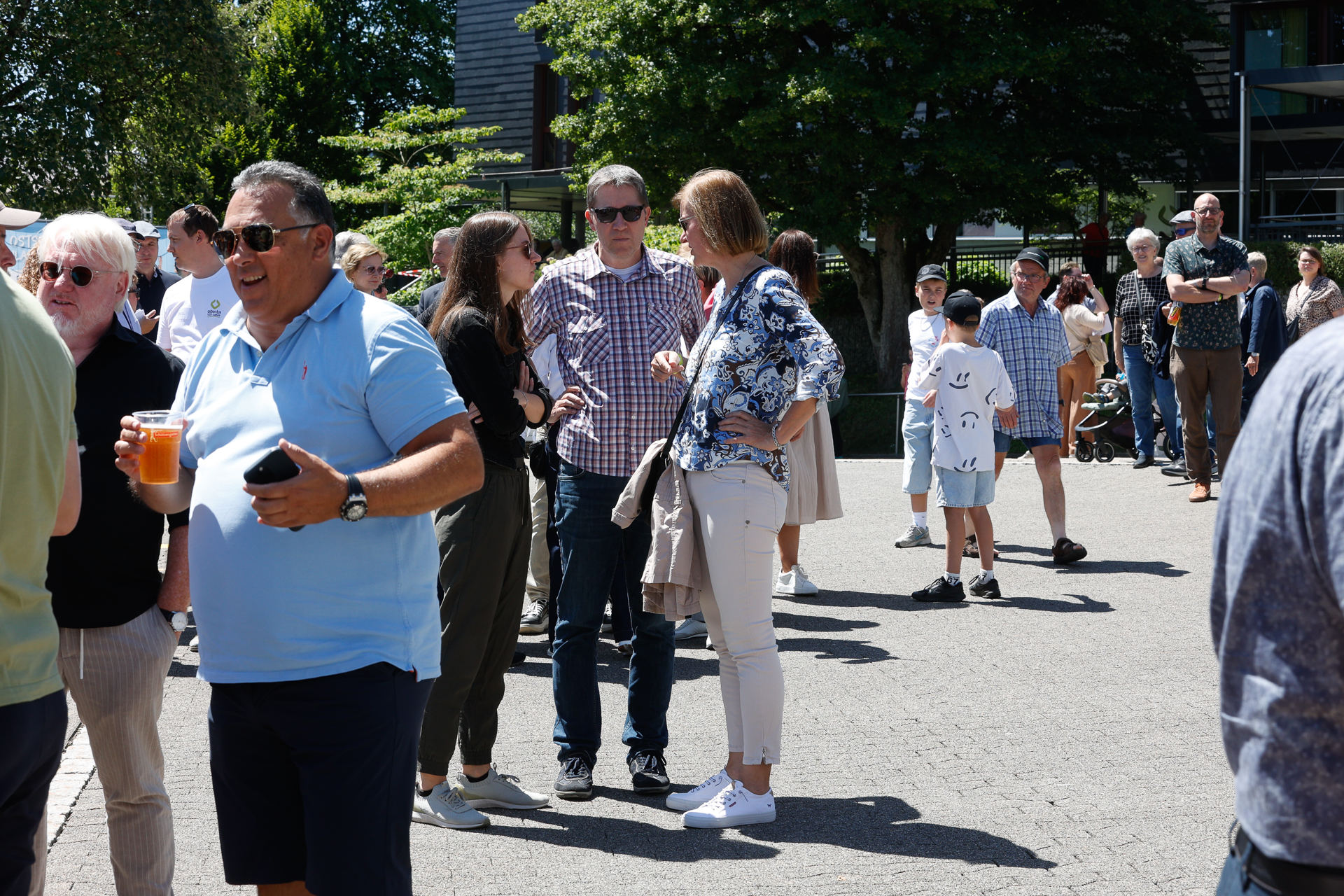 Eine Gruppe von Menschen steht draußen an einem sonnigen Ort; einige halten Getränke in der Hand und unterhalten sich, während andere in einer locker geformten Reihe warten. Im Hintergrund sind Bäume und Gebäude zu sehen.
