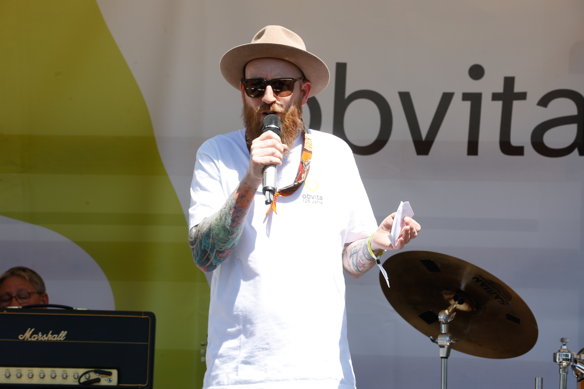 Ein Mann mit Bart und Tattoos, der eine Sonnenbrille, eine Mütze und ein weißes T-Shirt trägt, spricht auf einer Bühne im Freien in ein Mikrofon. Hinter ihm ist Musikausrüstung zu sehen, darunter ein Schlagzeug und ein Verstärker.