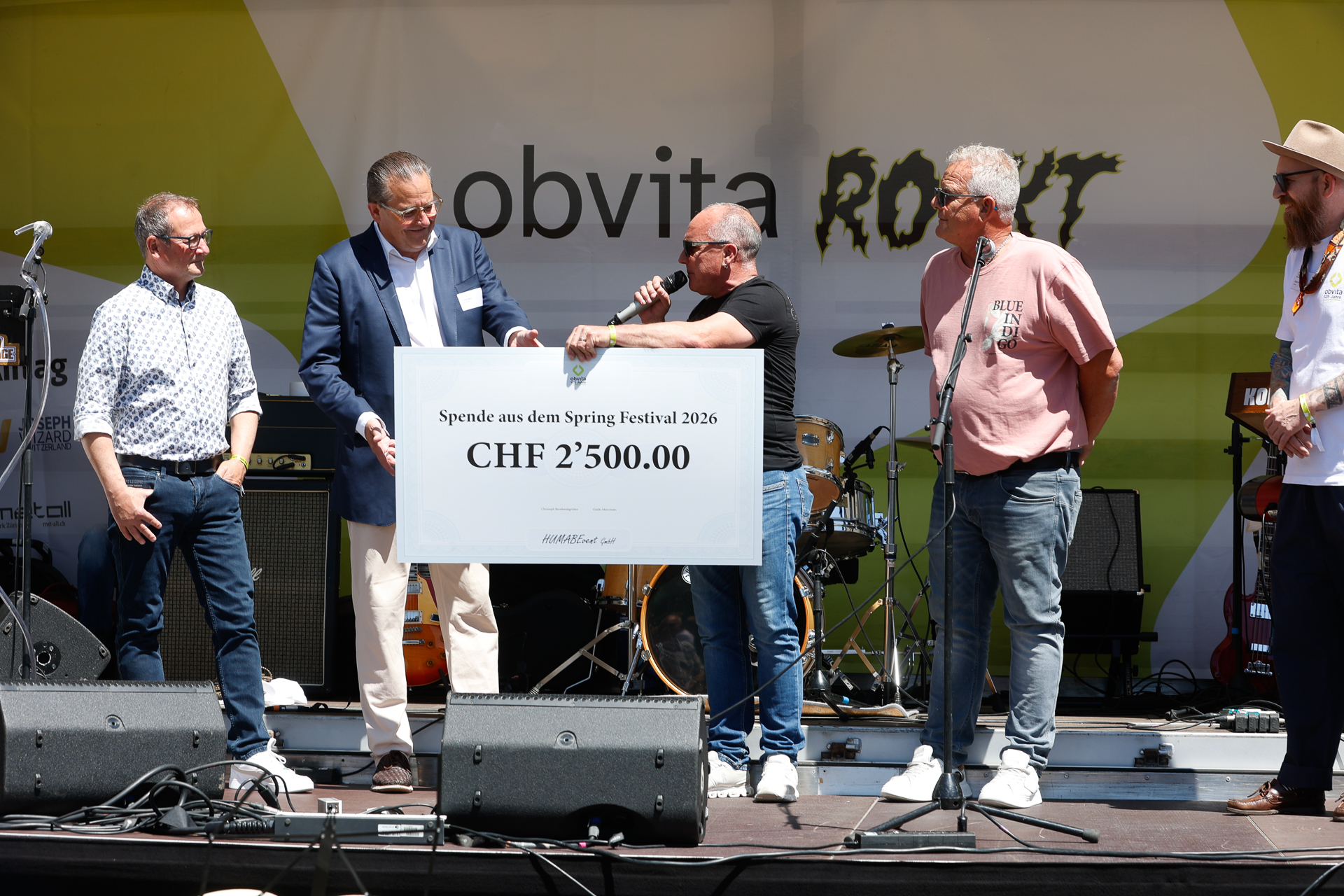 Fünf Männer stehen während einer Veranstaltung auf einer Bühne. Ein Mann spricht in ein Mikrofon, während er einen großen Spendenscheck über 2'500.00 CHF überreicht. Im Hintergrund sind Musikinstrumente und ein gelb-weißes Transparent zu sehen.