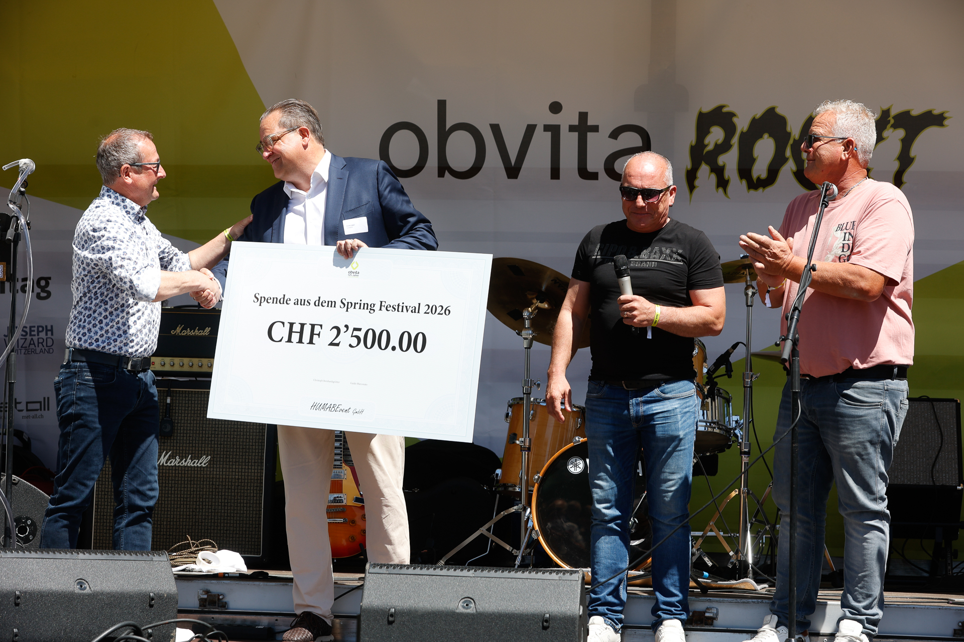 Vier Männer stehen auf der Bühne; einer schüttelt einem anderen die Hand, der einen großen Spendenscheck über 2.500 CHF in der Hand hält. Zwei weitere Männer stehen daneben, einer klatscht. Im Hintergrund sind Schlagzeuge und Mikrofone zu sehen.