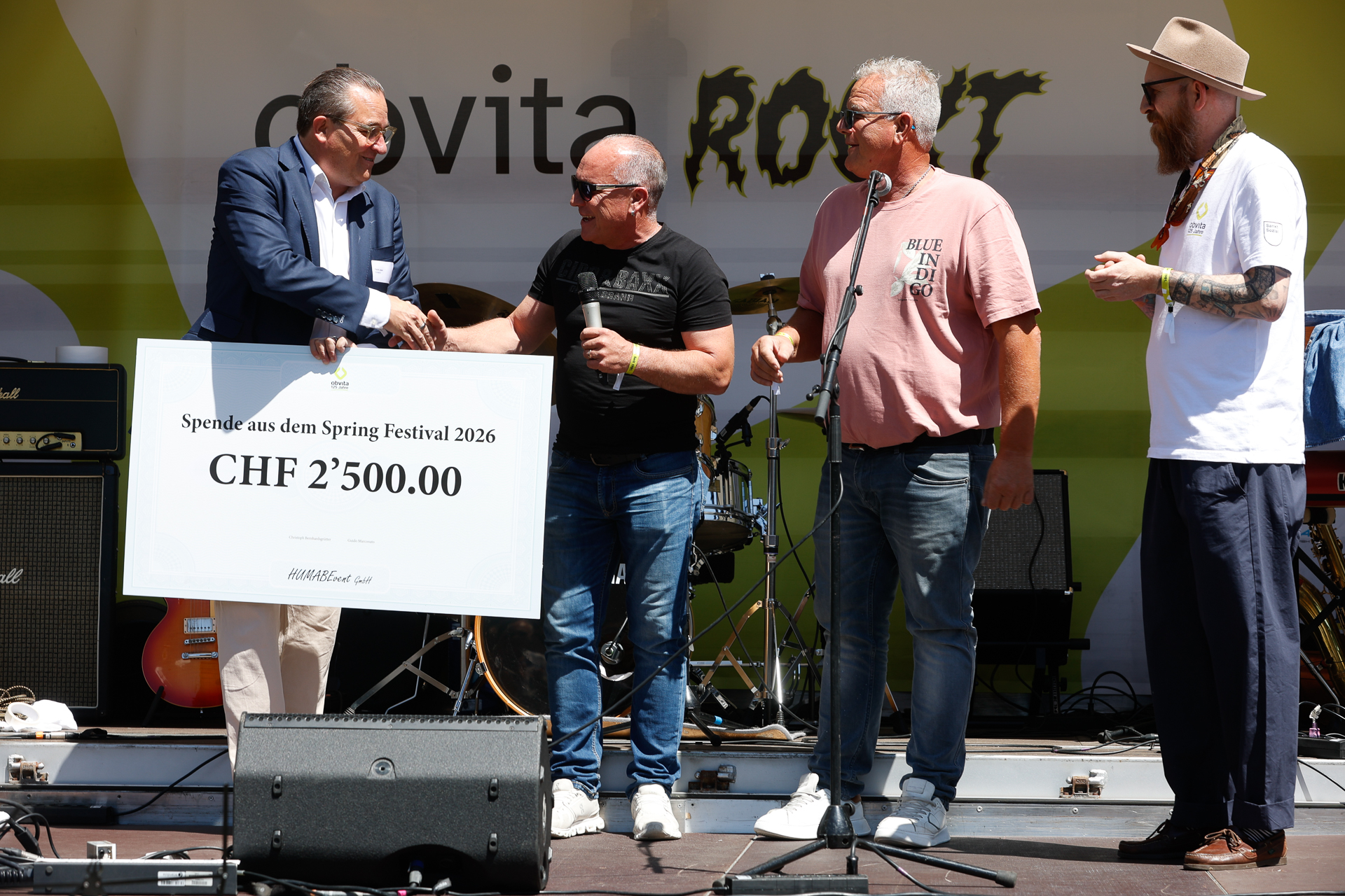 Vier Männer stehen auf der Bühne; einer überreicht einem anderen einen großen Scheck über 2'500 CHF. Im Hintergrund sind Musikinstrumente zu sehen, und hinter ihnen ist teilweise ein Festivalbanner zu erkennen. Die Szene wirkt festlich.