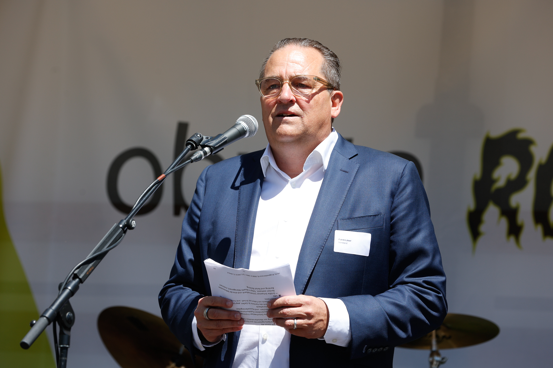 Ein Mann in einem marineblauen Anzug und einem weißen Hemd spricht in ein Mikrofon, während er einige Papiere in der Hand hält. Er steht im Freien vor einem unscharfen Hintergrund, auf dem undeutliche Schriftzüge und ein Schlagzeug zu erkennen sind.