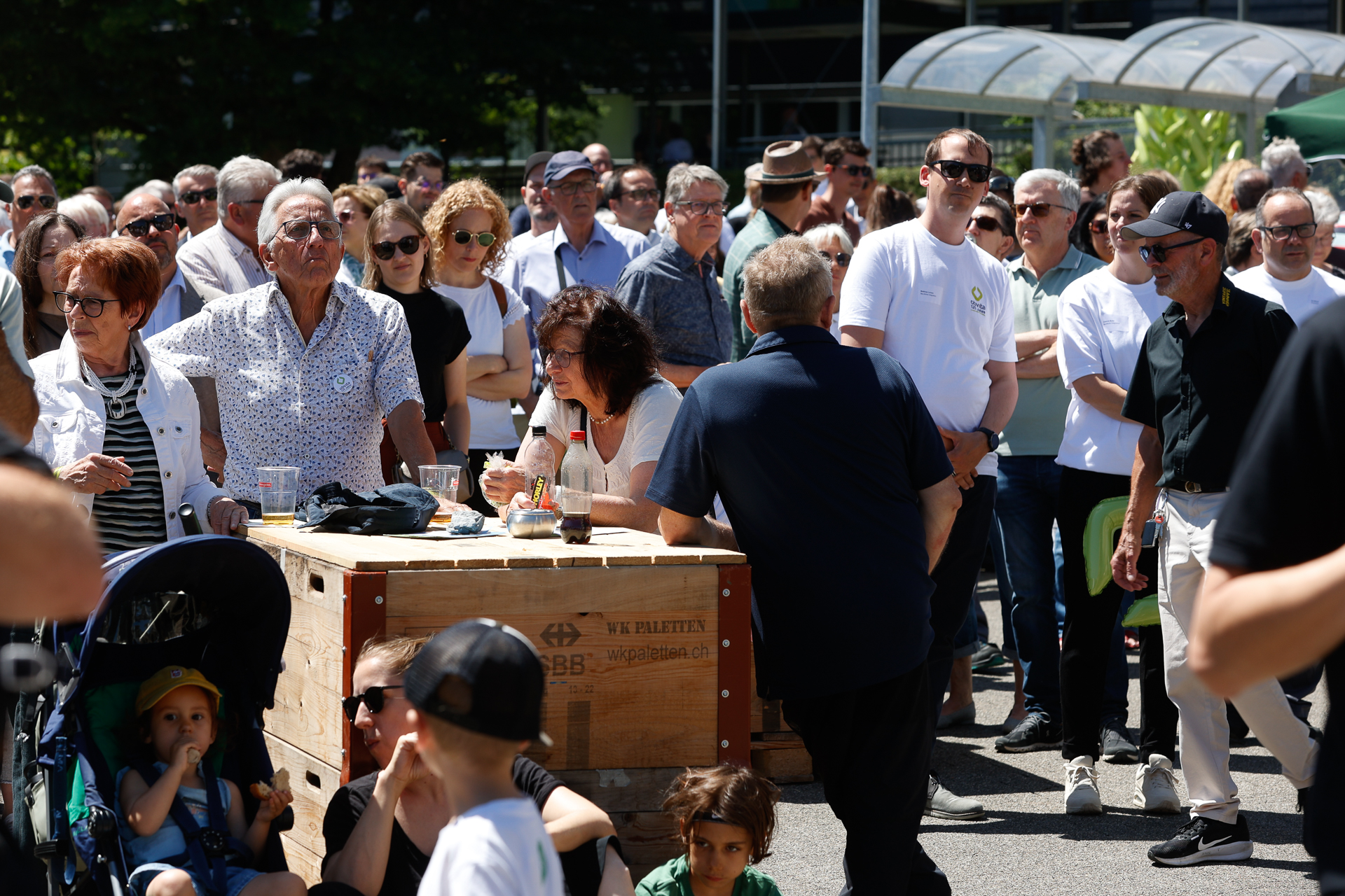 An einem sonnigen Tag versammelt sich eine große Gruppe von Menschen, darunter Kinder und Erwachsene, im Freien. Einige stehen, andere sitzen an einem Holztisch, und viele scheinen etwas außerhalb des Bildausschnitts zu beobachten oder zuzuhören.