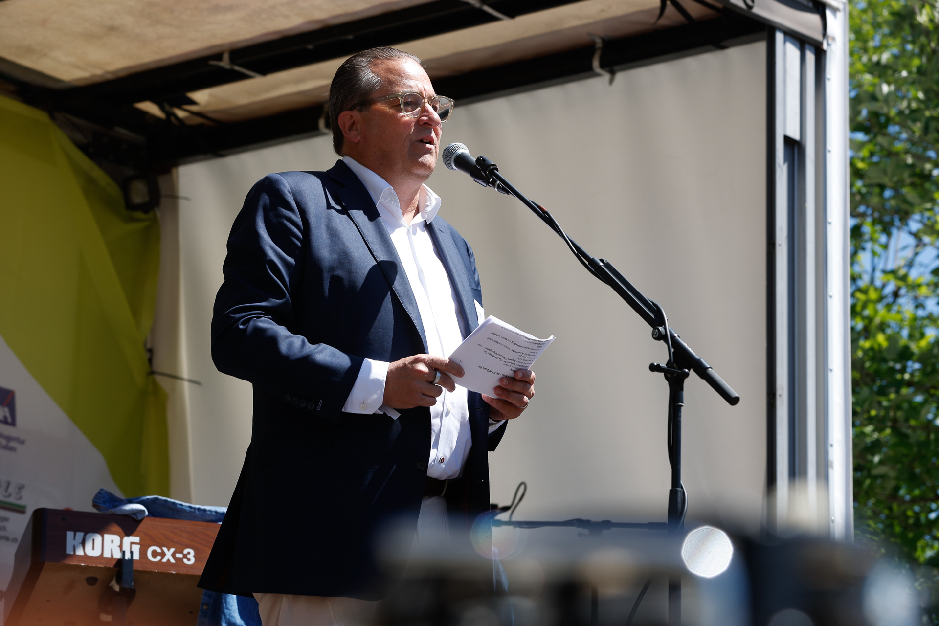Ein Mann in Anzug und Brille spricht auf einer Freiluftbühne in ein Mikrofon und hält dabei einige Blätter in der Hand. Sonnenlicht fällt auf sein Gesicht, und im Hintergrund sind Bäume zu sehen.