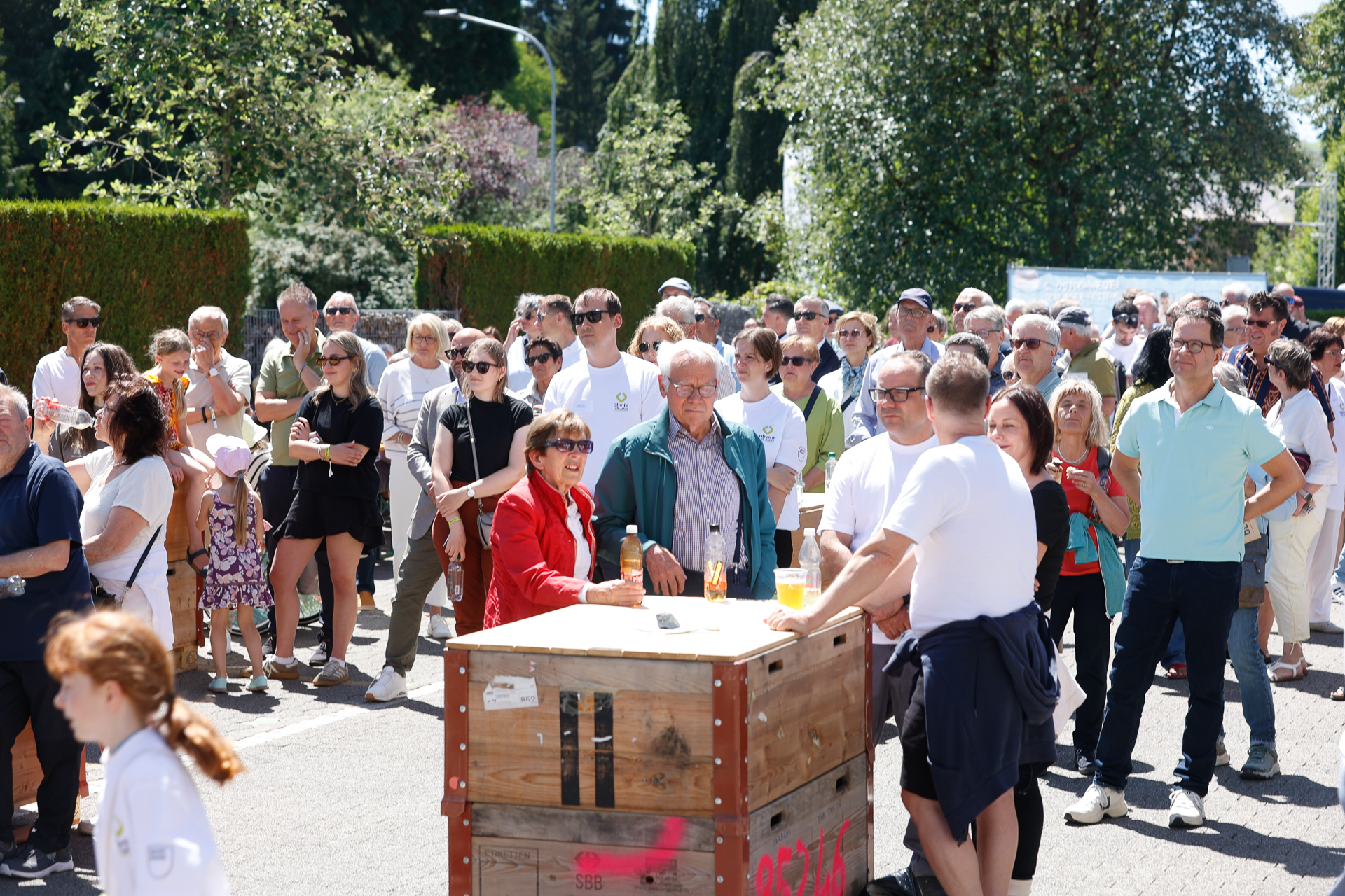 An einem sonnigen Tag versammelt sich draußen eine Menschenmenge; einige stehen um eine Holzkiste mit Getränken herum. Im Hintergrund sind Bäume und Grünflächen zu sehen, und die meisten Menschen tragen legeren Sommerkleidung und Sonnenbrillen.
