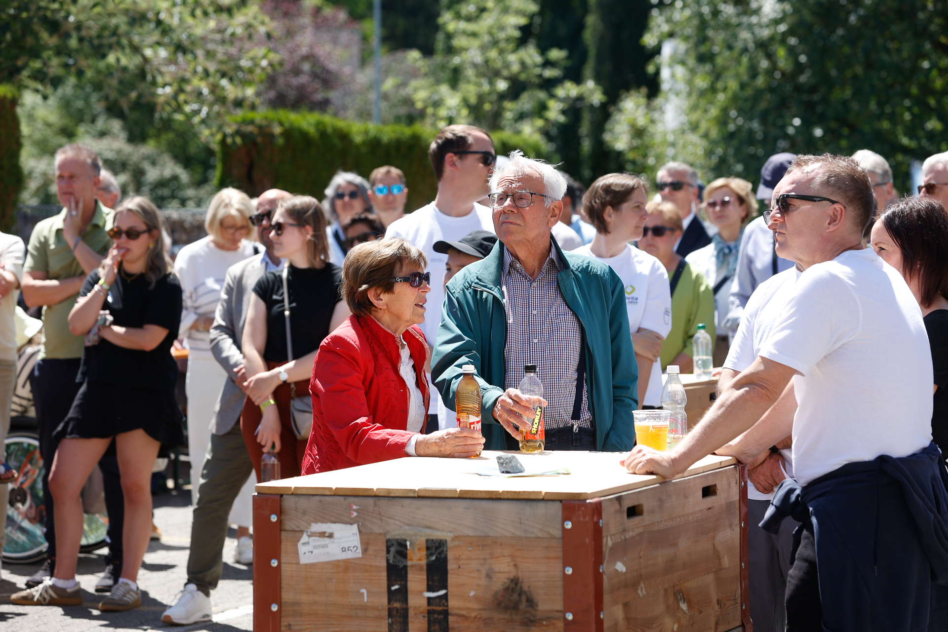 An einem sonnigen Tag versammelt sich eine Gruppe von Menschen im Freien; viele tragen Freizeitkleidung und Sonnenbrillen. Ein älterer Mann und eine ältere Frau stehen an einem Holztisch mit Getränken, umgeben von anderen, die etwas außerhalb des Bildausschnitts beobachten.