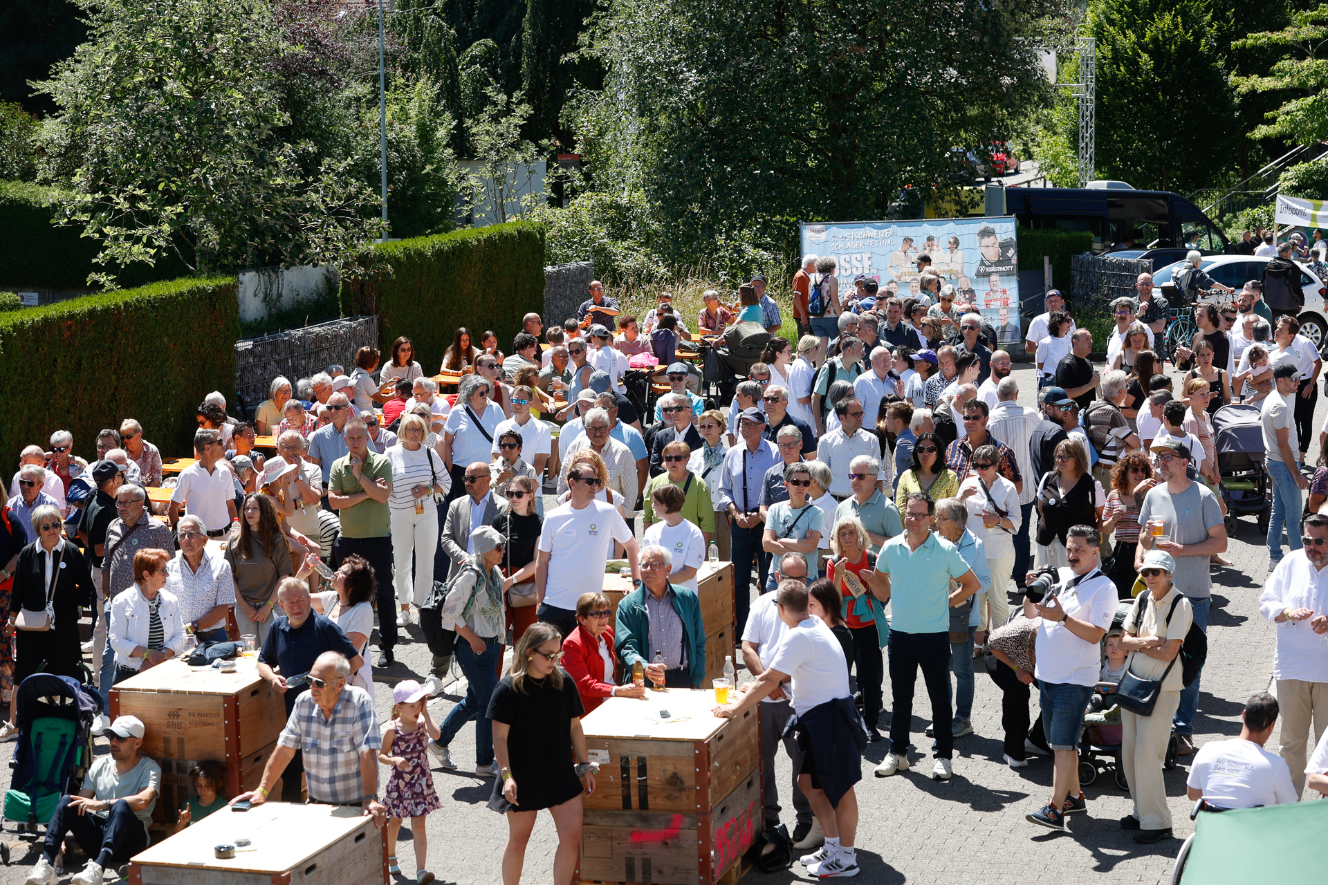 An einem sonnigen Tag versammelt sich eine große Gruppe von Menschen im Freien; sie stehen um Holztische herum und unterhalten sich. Einige tragen Sonnenbrillen und Hüte. Im Hintergrund sind Bäume, Hecken und Banner zu sehen.