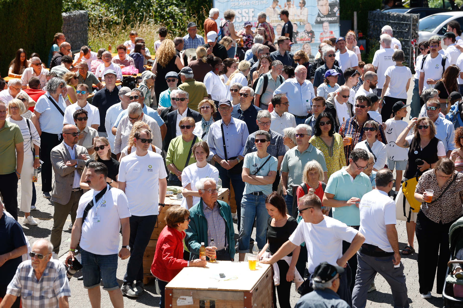 An einem sonnigen Tag versammelte sich draußen eine große Menschenmenge; viele trugen Sonnenbrillen und legeren Sommerkleidung, einige hielten Getränke in der Hand und unterhielten sich, im Hintergrund waren Tische und Grünflächen zu sehen.