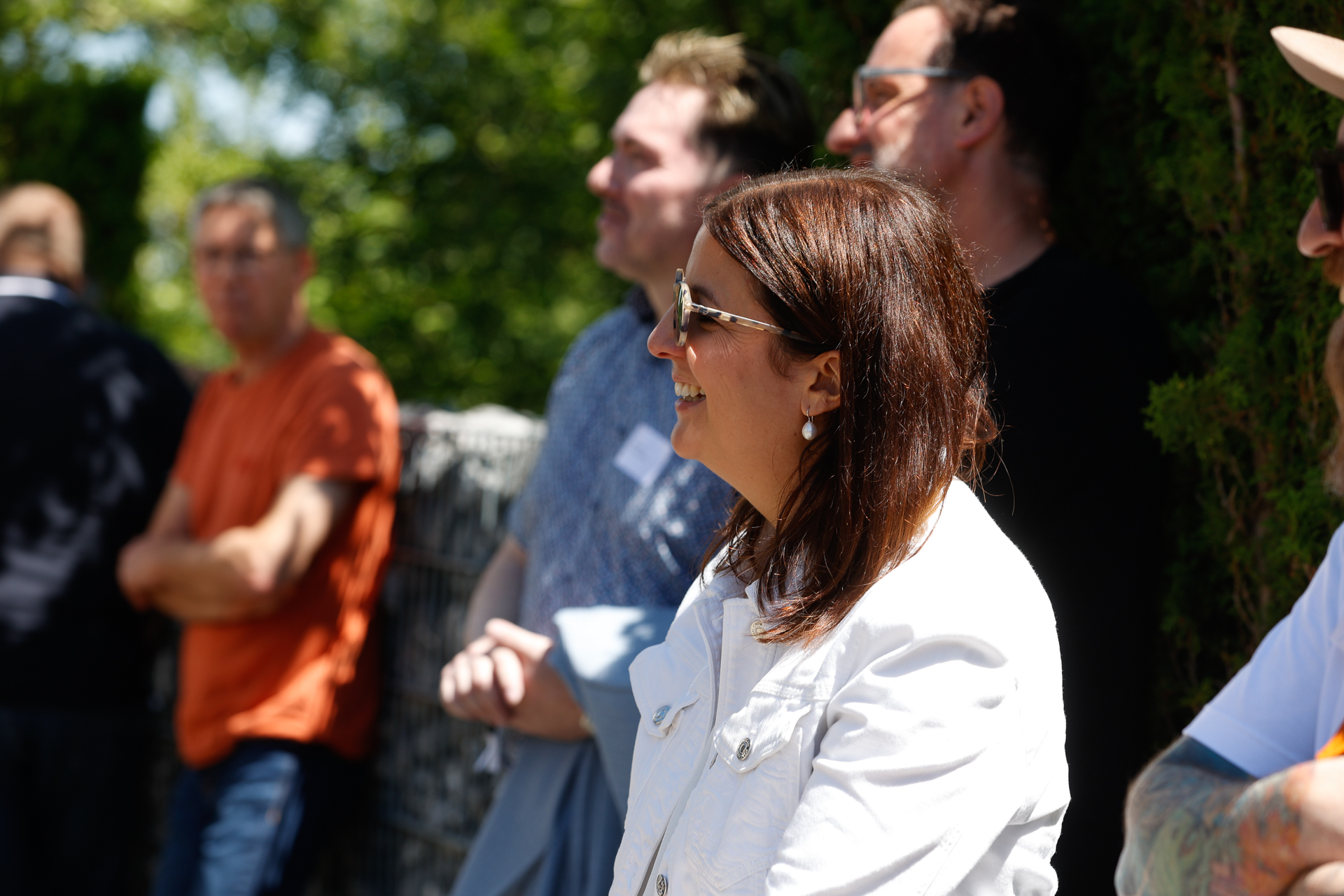Eine Frau in einer weißen Jacke und mit Sonnenbrille lächelt im Freien inmitten einer Gruppe von Menschen, im Hintergrund sind Bäume und Sonnenlicht zu sehen. Andere Menschen stehen da und beobachten etwas außerhalb des Bildausschnitts.