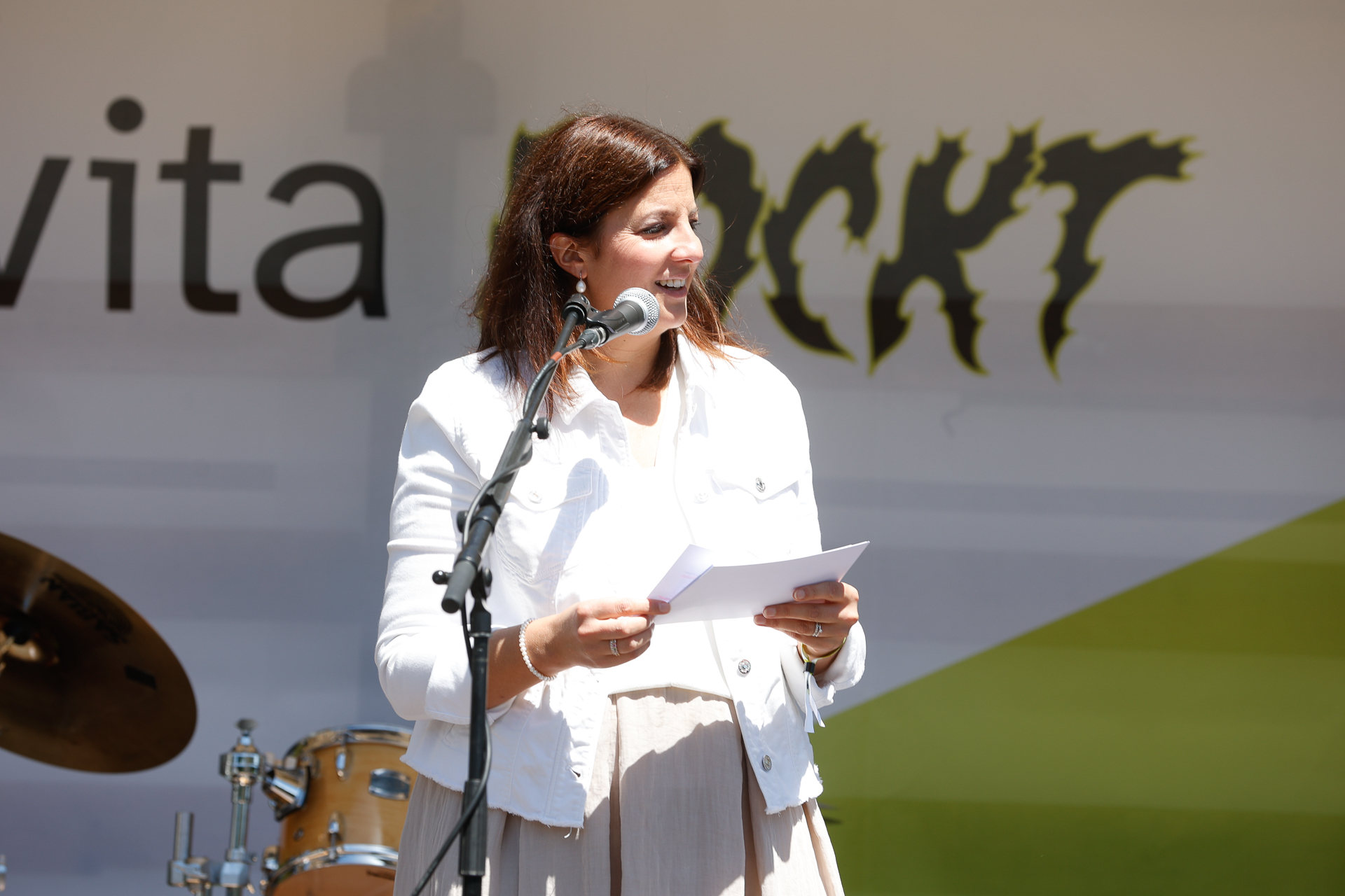 Eine Frau in einer weißen Jacke steht am Mikrofon, hält Papiere in der Hand und hält bei einer Veranstaltung im Freien eine Rede. Im Hintergrund sind ein Schlagzeug und ein teilweise sichtbares Veranstaltungsbanner zu sehen.
