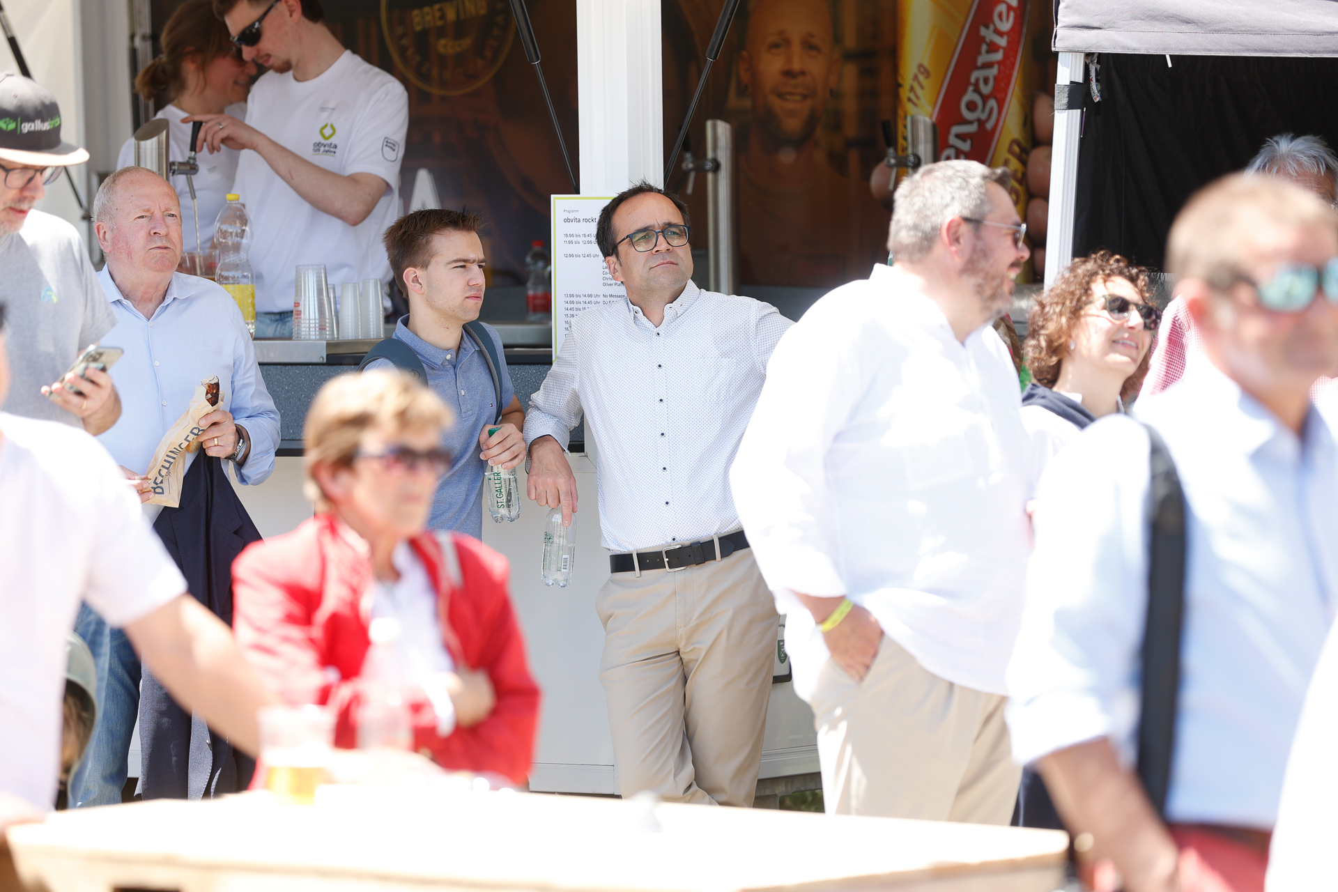 Eine Gruppe von Menschen, überwiegend Männer in weißen Hemden und mit Sonnenbrillen, steht bei Tageslicht im Freien; einige halten Wasserflaschen in der Hand, während sie interessiert etwas außerhalb des Bildausschnitts beobachten.