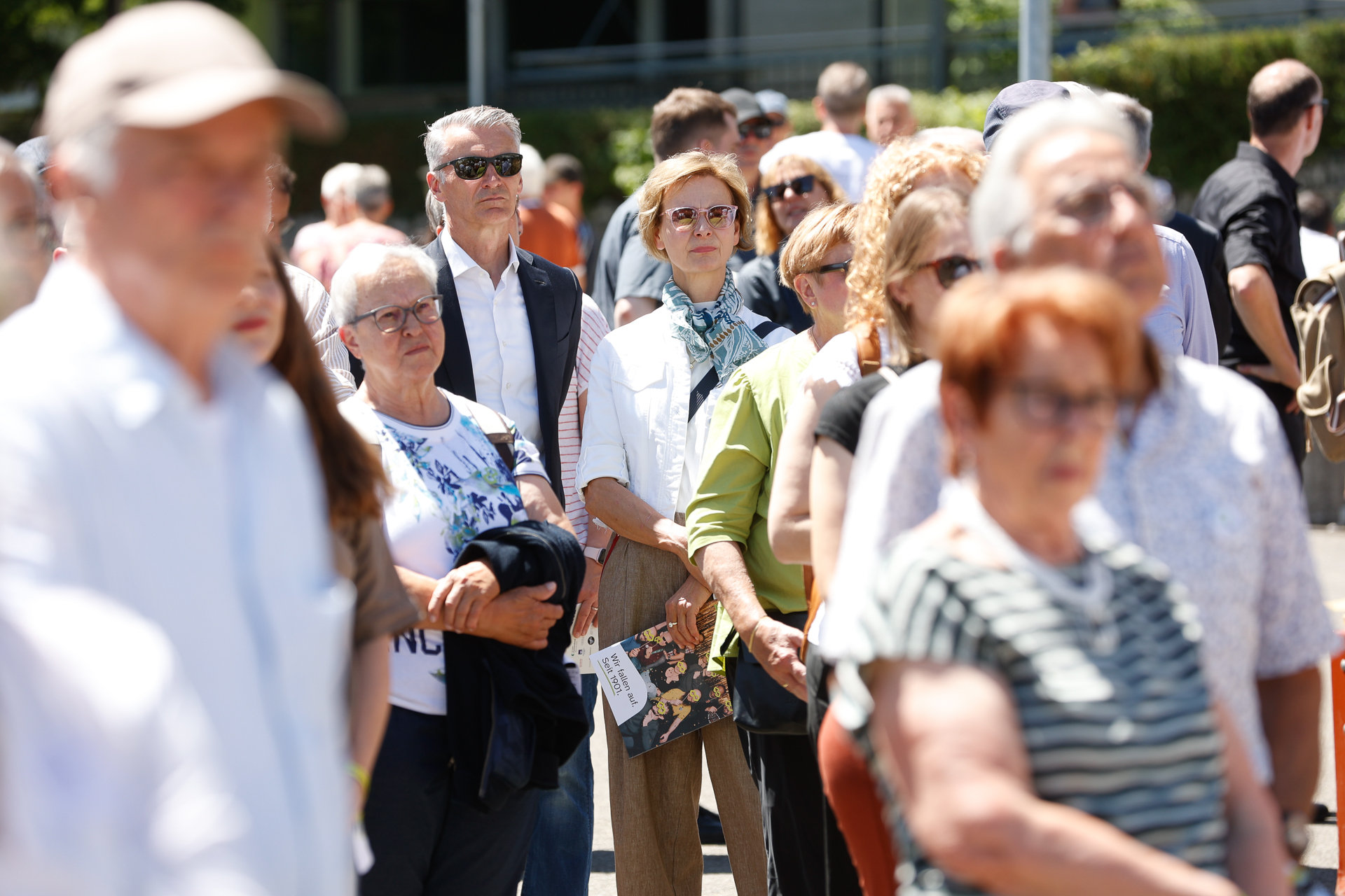 Eine Gruppe Erwachsener steht im Freien im Sonnenlicht, einige tragen Sonnenbrillen. Viele wirken aufmerksam oder nachdenklich; in der Mitte steht eine Frau mit Schal und Sonnenbrille. Im Vordergrund und Hintergrund sind mehrere Personen unscharf abgebildet.