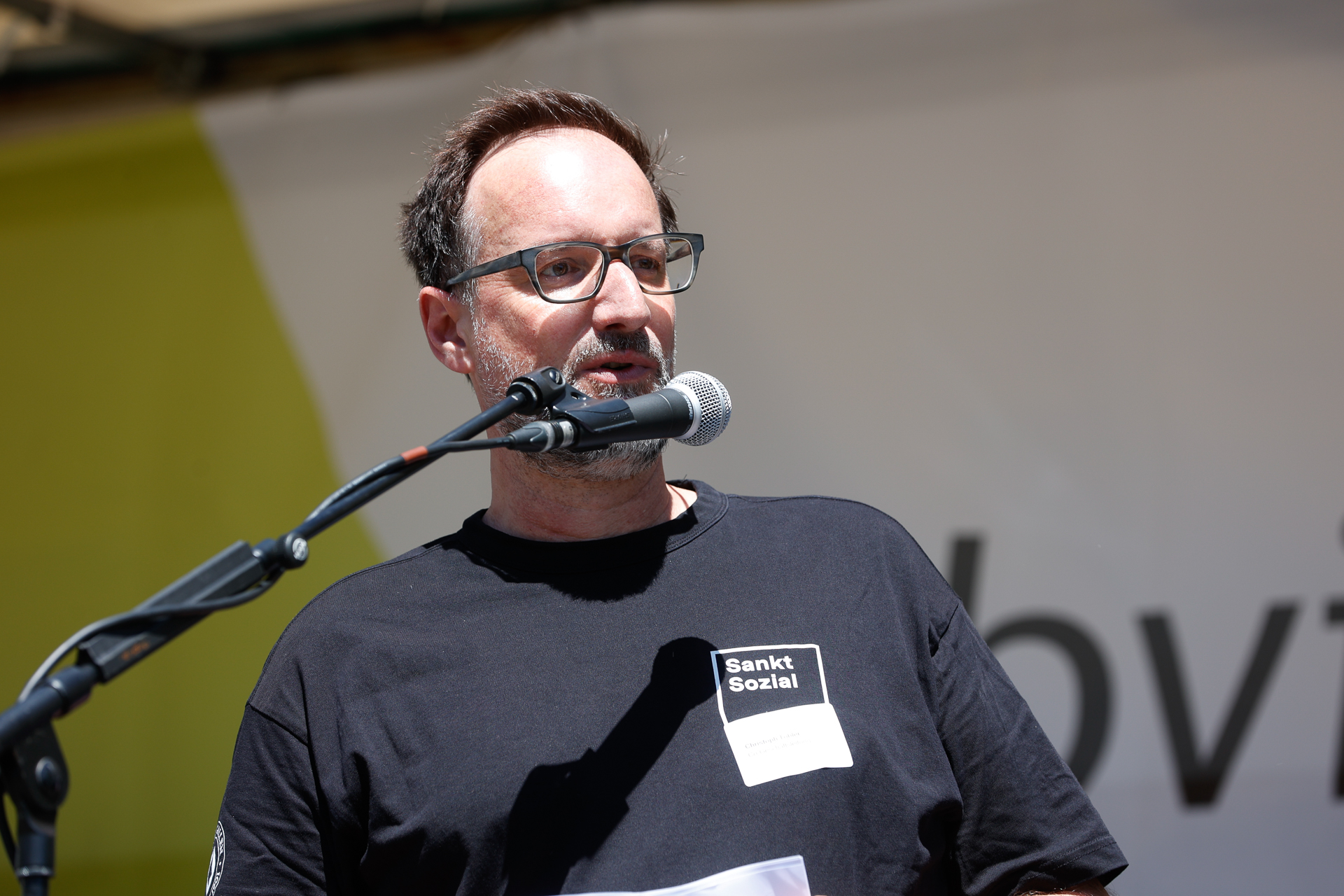 Ein Mann mit Brille und einem schwarzen „Sankt Sozial“-T-Shirt spricht bei einer Veranstaltung im Freien in ein Mikrofon und hält dabei einige Blätter in der Hand. Der Hintergrund ist in Gelb- und Weißtönen verschwommen.