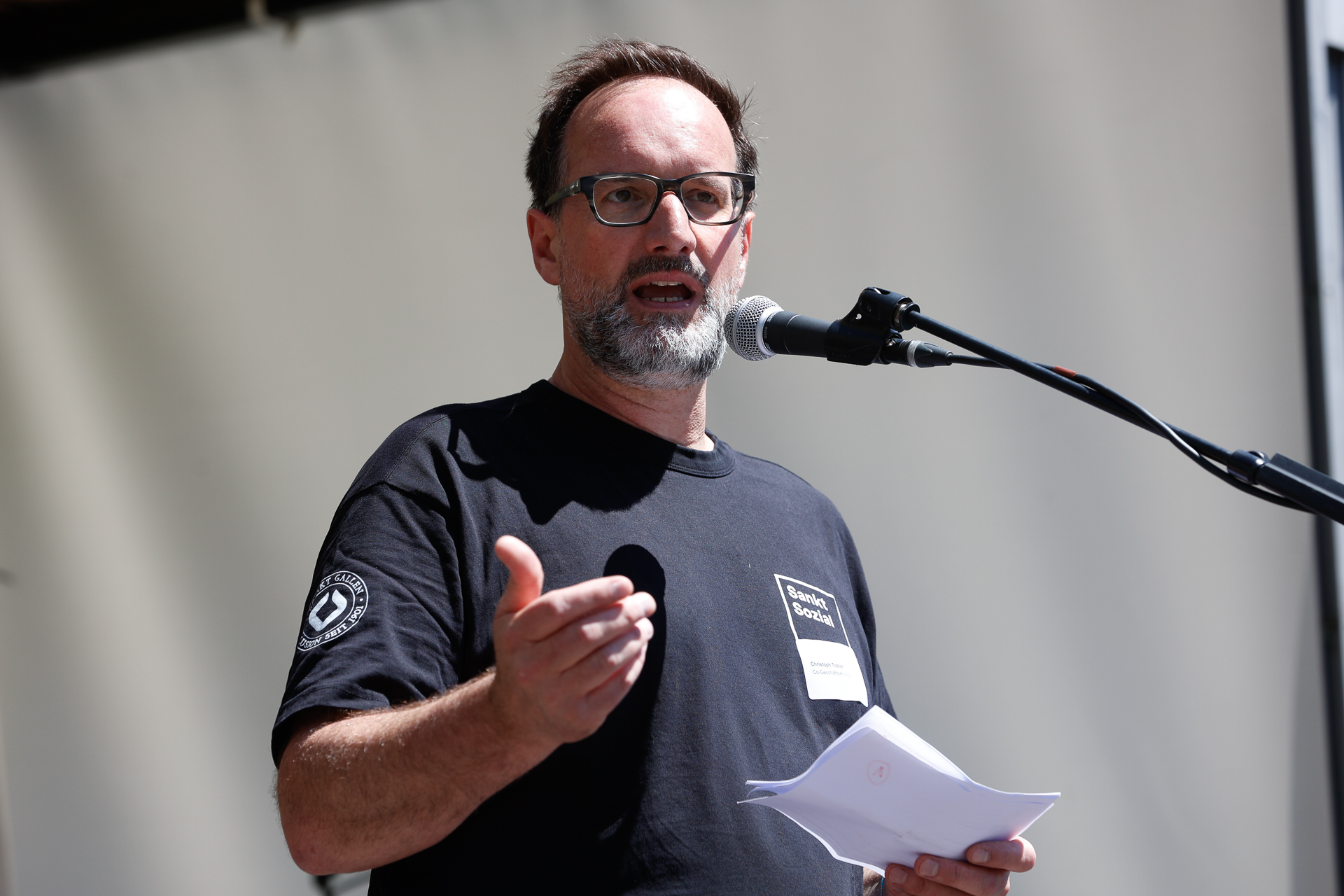 Ein Mann mit Brille und Bart spricht in ein Mikrofon, während er Papiere in der Hand hält und mit einer Hand gestikuliert. Er trägt ein schwarzes T-Shirt und steht vor einem schlichten, hellen Hintergrund.