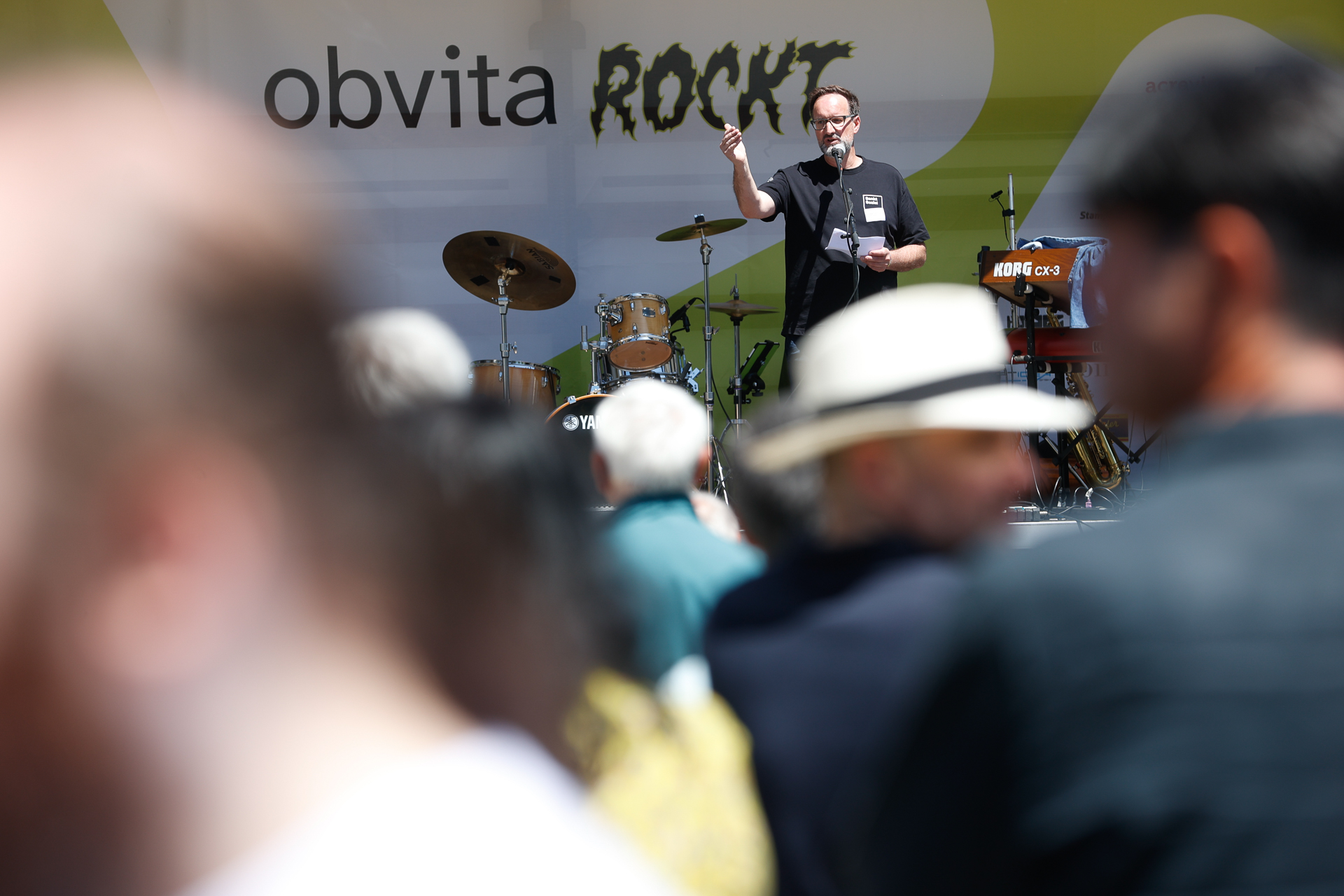 Eine Person steht auf einer Bühne im Freien und spricht in ein Mikrofon, hinter ihr stehen Musikinstrumente. Die Menschenmenge im Vordergrund ist leicht unscharf. Auf einem Banner im Hintergrund steht „obvita ROCKT“.