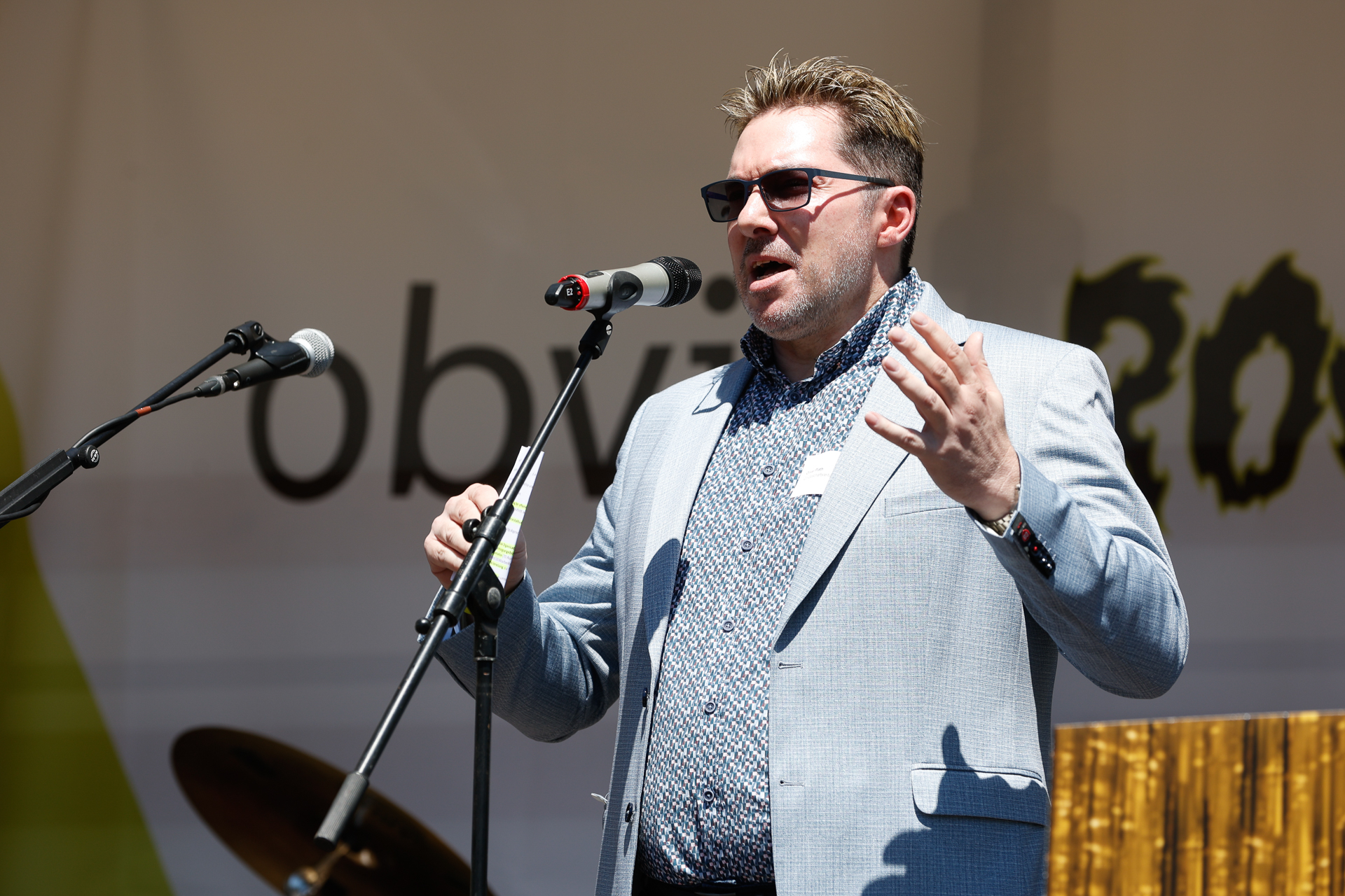 Ein Mann mit Sonnenbrille und grauem Blazer hält bei einer Veranstaltung im Freien eine leidenschaftliche Rede, während er vor den Mikrofonen auf einer Bühne mit verziertem Hintergrund steht.