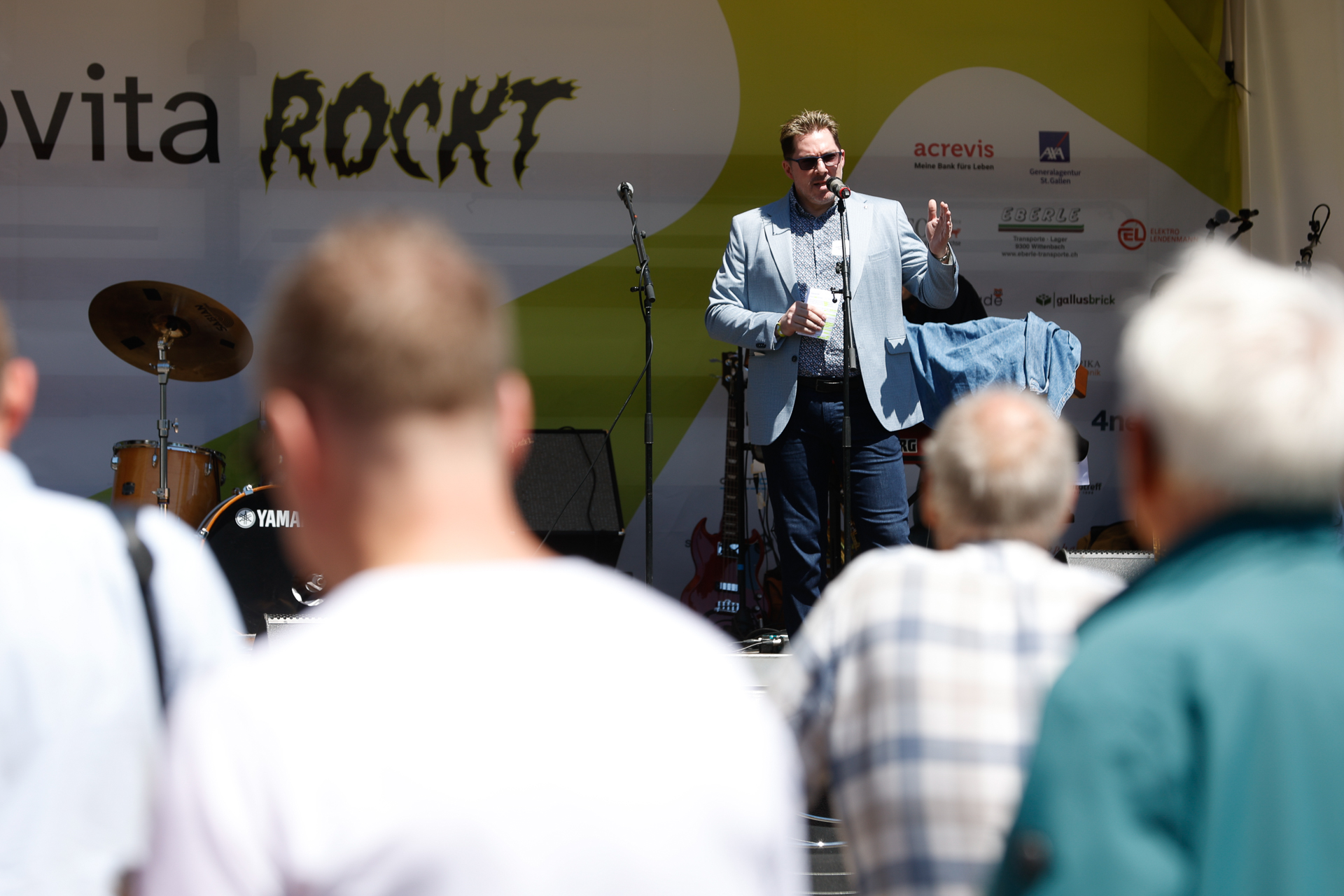 Ein Mann in einem blauen Blazer spricht auf einer Freiluftbühne in ein Mikrofon und blickt dabei ins Publikum. Hinter ihm sind Musikinstrumente zu sehen, und auf der Bühnenkulisse steht „ROCKT“. Im Vordergrund schauen mehrere Personen zu.