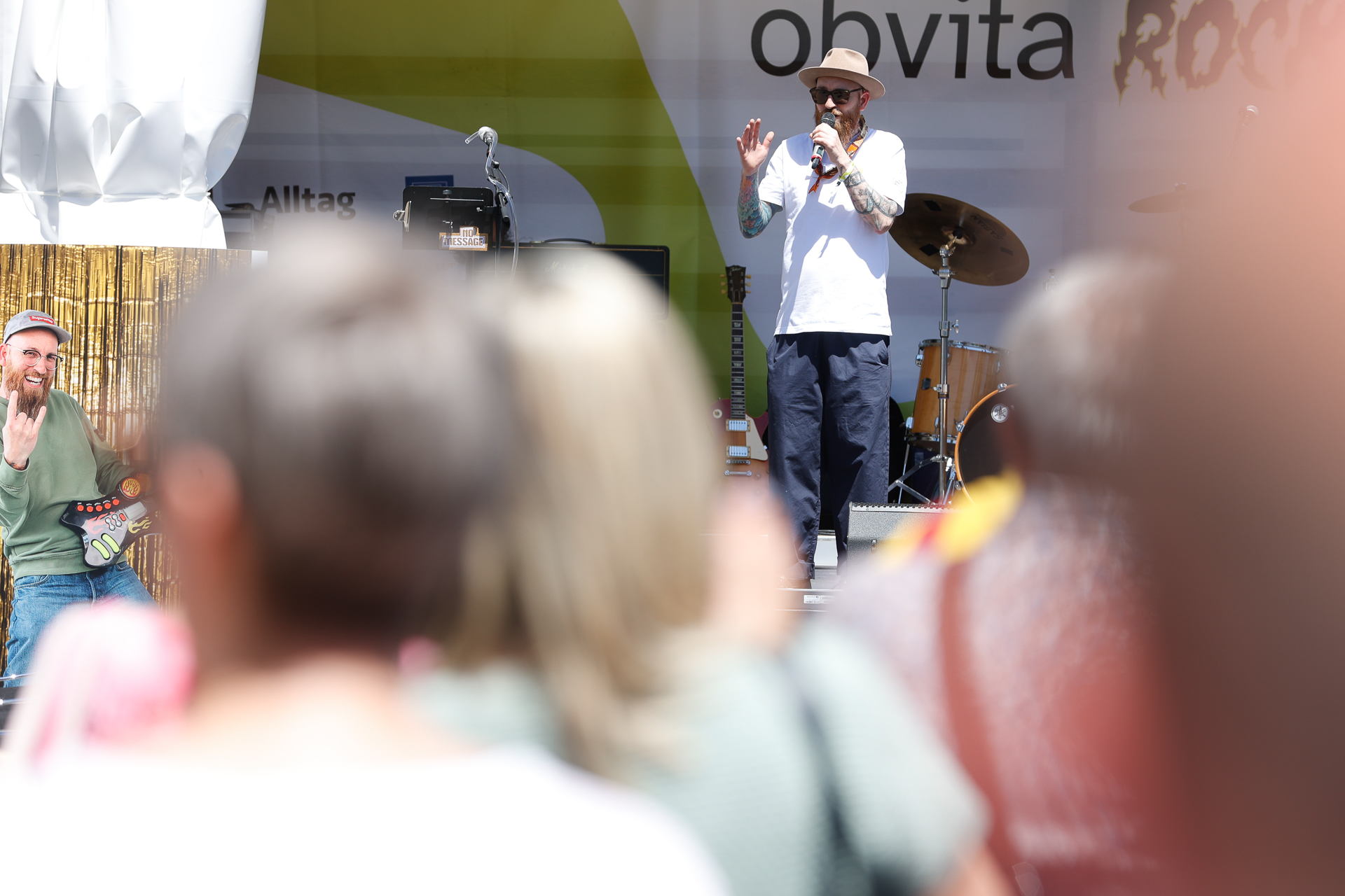 Ein Mann mit Hut und Sonnenbrille spricht auf einer Freiluftbühne vor Musikinstrumenten in ein Mikrofon, während unscharf abgebildete Zuschauer ihm zusehen.