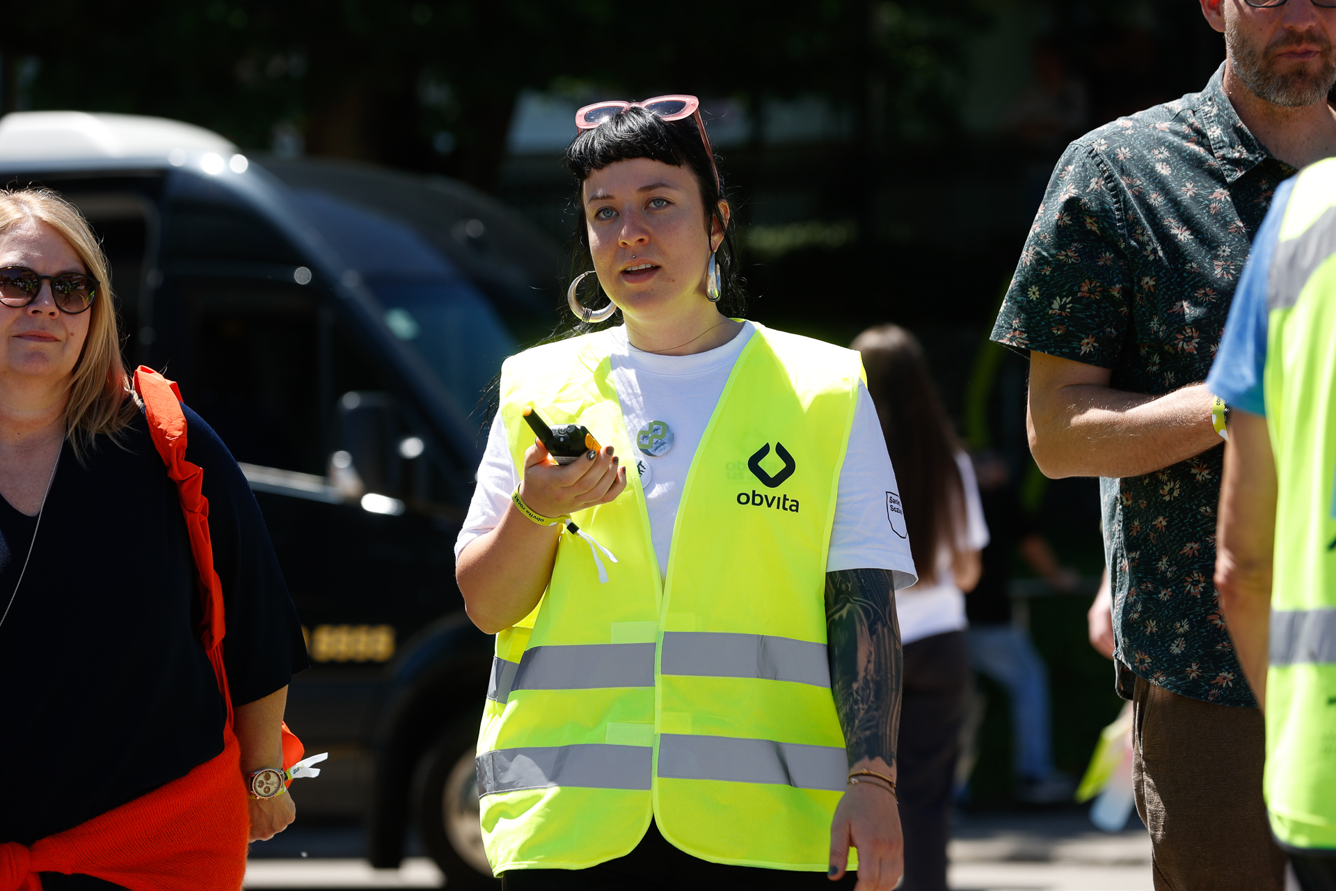 Eine Frau, die eine leuchtend gelbe Sicherheitsweste mit der Aufschrift „obvita“ trägt, hält ein Funkgerät in der Hand. Sie steht im Freien zusammen mit anderen Personen, darunter ein Mann und eine weitere Frau; alle scheinen an einer organisierten Veranstaltung teilzunehmen.