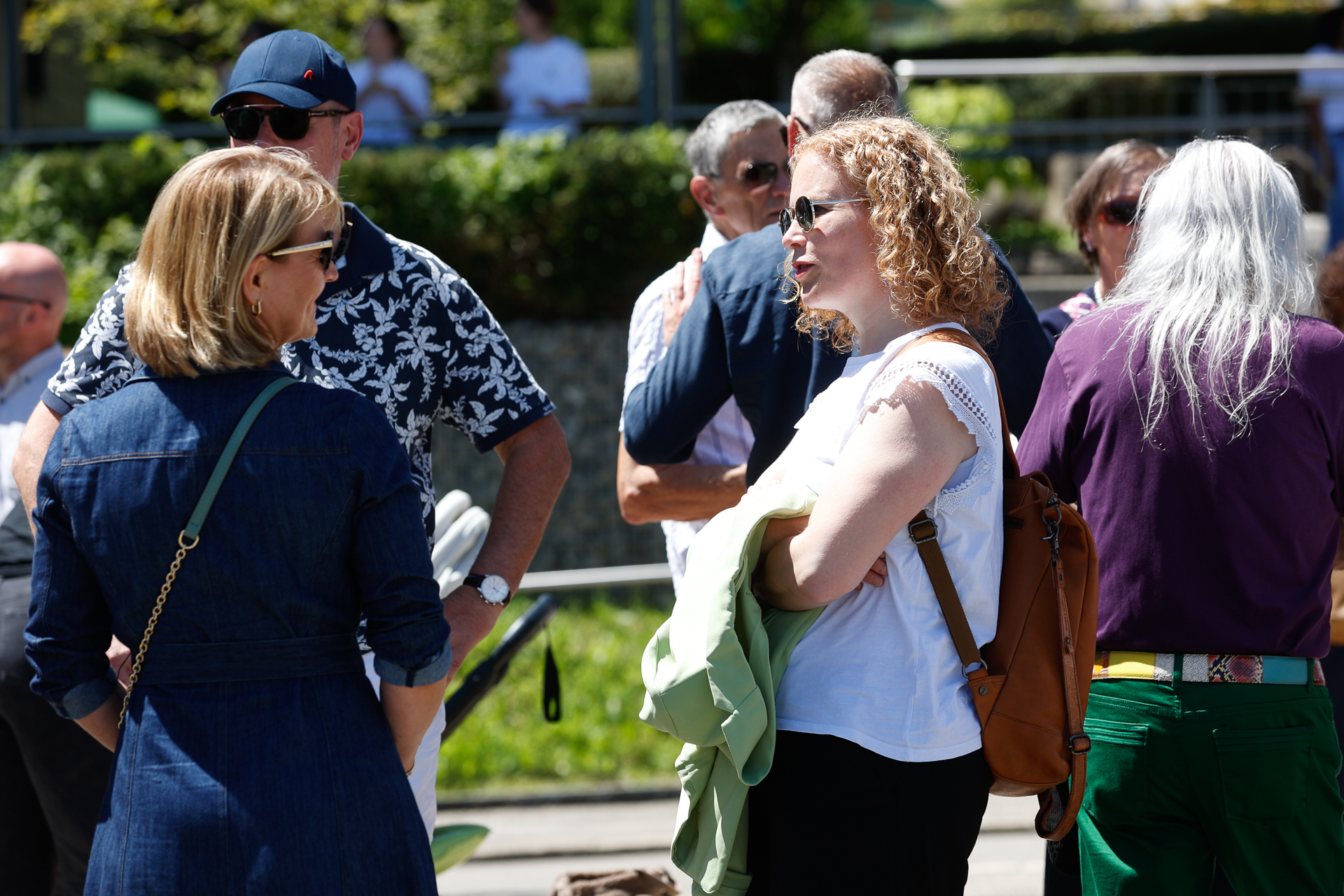 Eine Gruppe von Menschen steht bei sonnigem Wetter im Freien, unterhält sich und tauscht sich aus. Einige tragen Sonnenbrillen und leichte Kleidung. Im Hintergrund sind grüne Bäume und verschwommene Gestalten in weißen Hemden zu sehen.