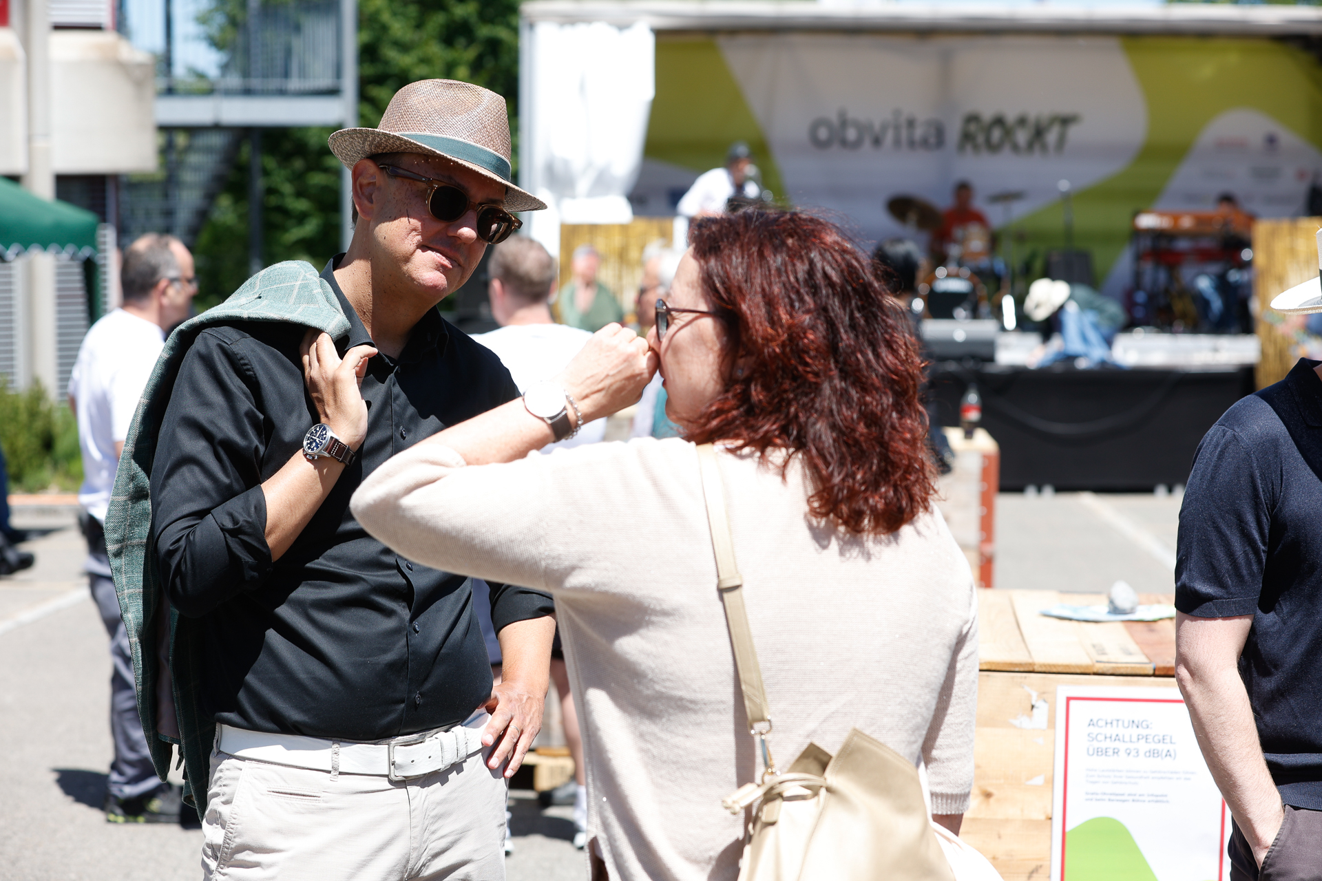 Ein Mann mit Sonnenbrille und Hut unterhält sich bei einer Veranstaltung im Freien mit einer Frau; im Hintergrund sind eine Bühne und ein Banner mit der Aufschrift „obvita ROCKT“ zu sehen. In der sonnigen Kulisse sind weitere Personen und Stände zu erkennen.