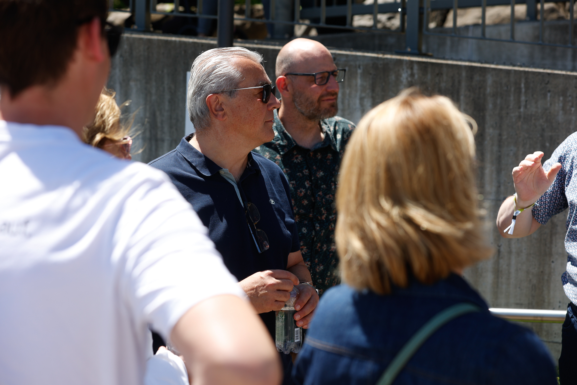 Eine Gruppe Erwachsener steht draußen im Sonnenlicht und unterhält sich. Ein Mann mit Sonnenbrille und dunklem Hemd hält eine Dose in der Hand. Andere, sowohl Männer als auch Frauen, sind teilweise zu sehen, einige stehen mit dem Rücken zur Kamera. Im Hintergrund ist eine Betonmauer zu erkennen.