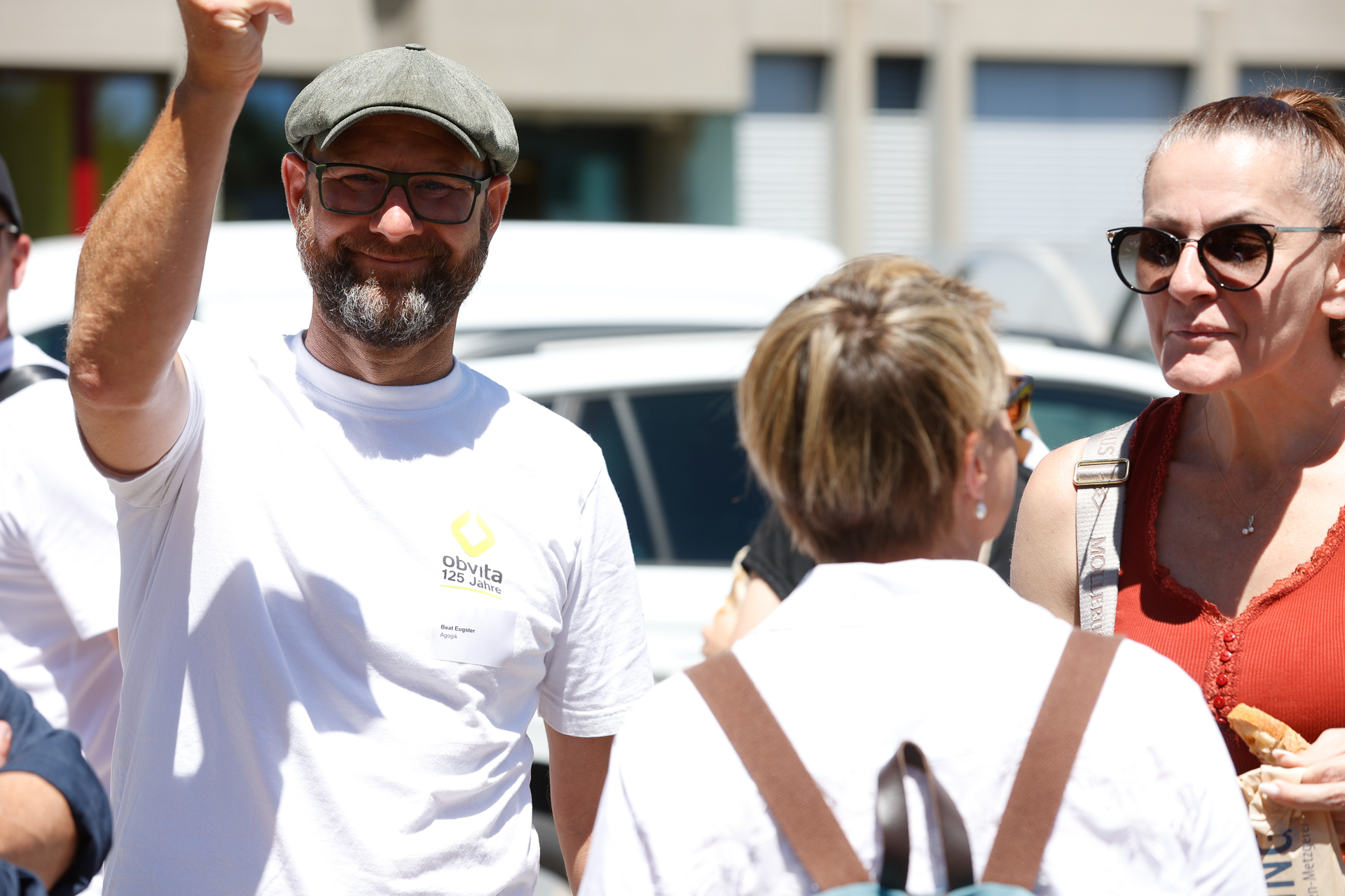 Ein Mann in einem weißen T-Shirt und einer Schirmmütze winkt und lächelt in die Kamera, während er an einem sonnigen Tag im Freien mit zwei Frauen steht – eine von ihnen trägt eine Sonnenbrille und ein orangefarbenes Oberteil – inmitten einer kleinen Gruppe von Menschen.