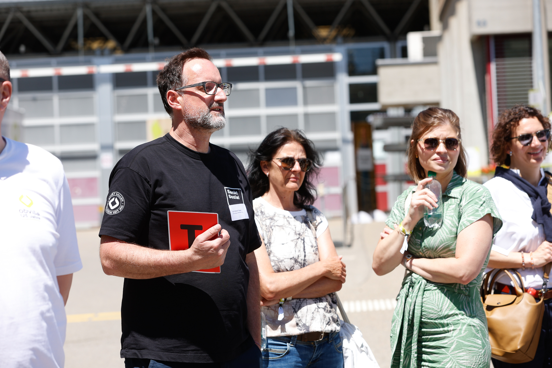 An einem sonnigen Tag steht eine Gruppe von Menschen im Freien. Ein Mann in einem schwarzen Hemd hält eine rote Mappe in der Hand und trägt eine Brille, während die anderen um ihn herum in legerer Sommerkleidung und mit Sonnenbrillen zuhören. Im Hintergrund ist ein Industriegebäude zu sehen.