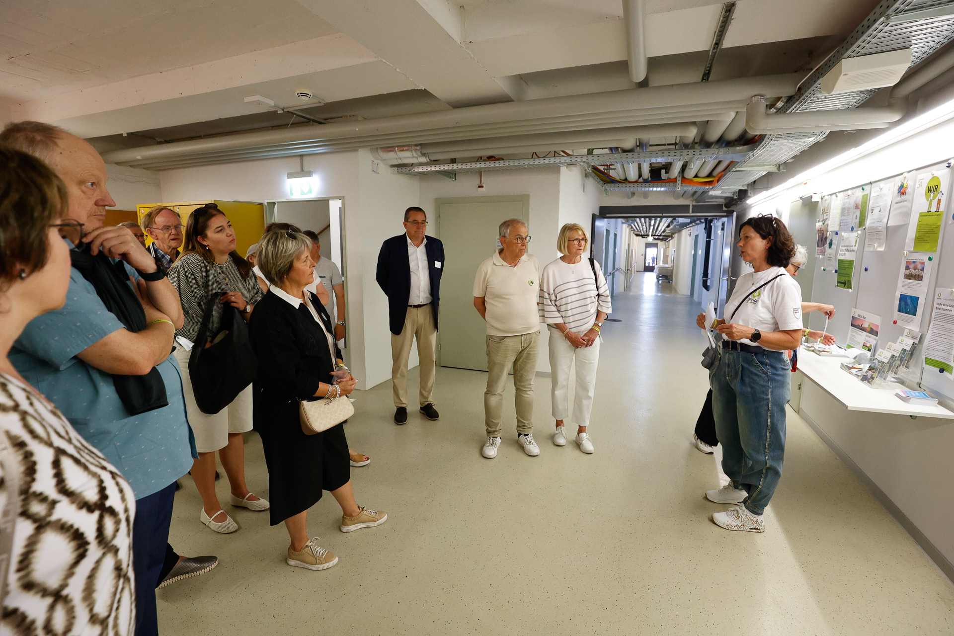 Eine Gruppe von Menschen steht in einem Flur und hört einer Frau zu, die neben einer Ausstellung aus Plakaten und Papieren an der Wand spricht. Der Schauplatz scheint eine Führung oder eine Informationsveranstaltung in einem institutionellen Gebäude zu sein.
