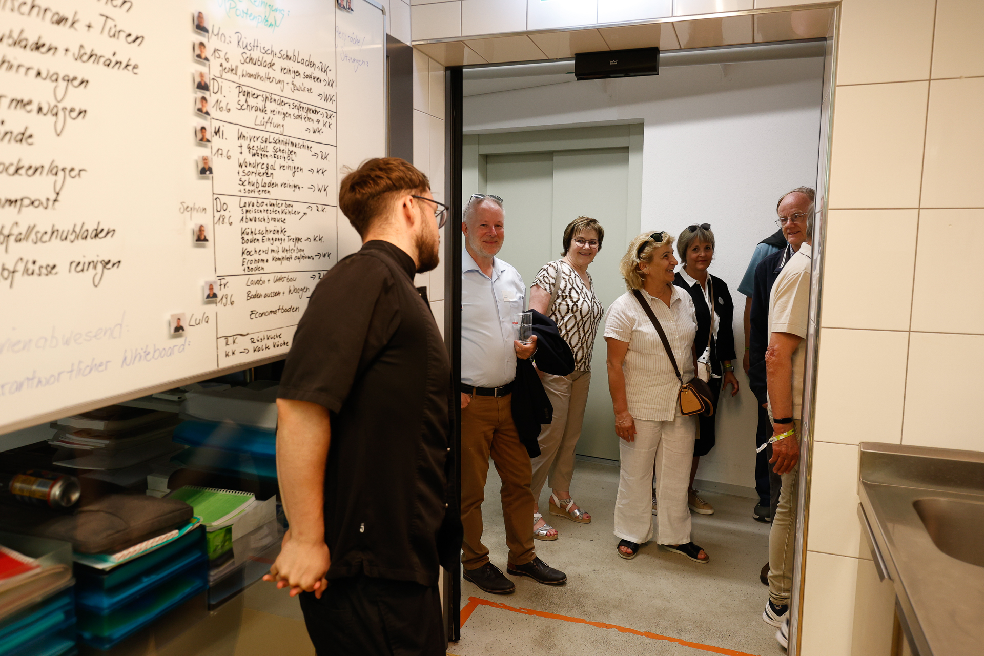 Eine Gruppe von sechs Erwachsenen steht gemeinsam in einer Küchentür, einige lächeln. Ein Mann in schwarzer Kleidung steht daneben. An der linken Wand ist eine Weißwandtafel mit Notizen auf Deutsch zu sehen. Die Küche hat geflieste Wände und Regale.