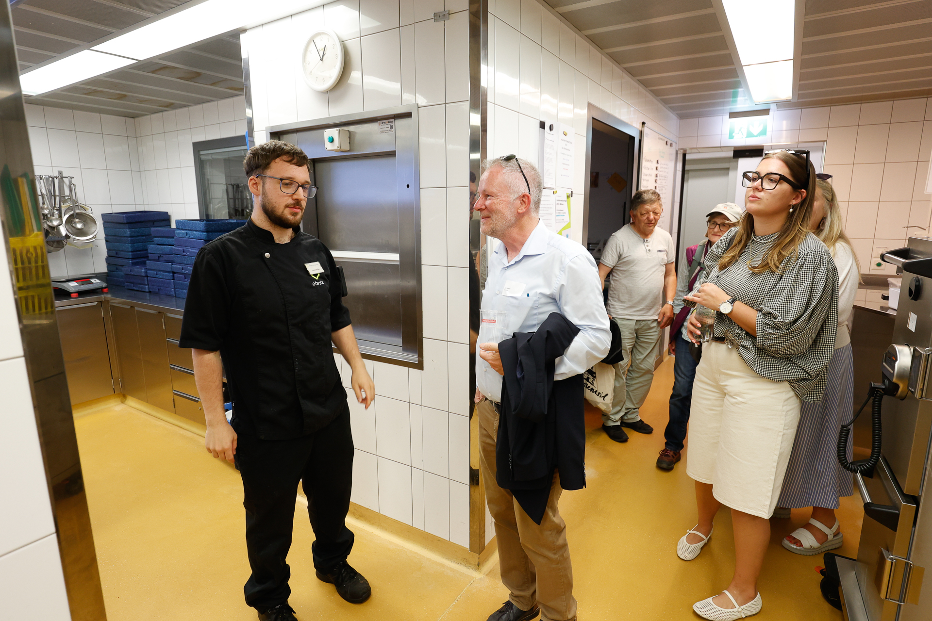 Ein Koch in schwarzer Uniform unterhält sich mit einer Gruppe von Besuchern, die eine saubere, moderne Küche mit weiß gefliesten Wänden und gelbem Boden besichtigen. Einige Besucher hören aufmerksam zu, an der Wand hängen Küchenutensilien.
