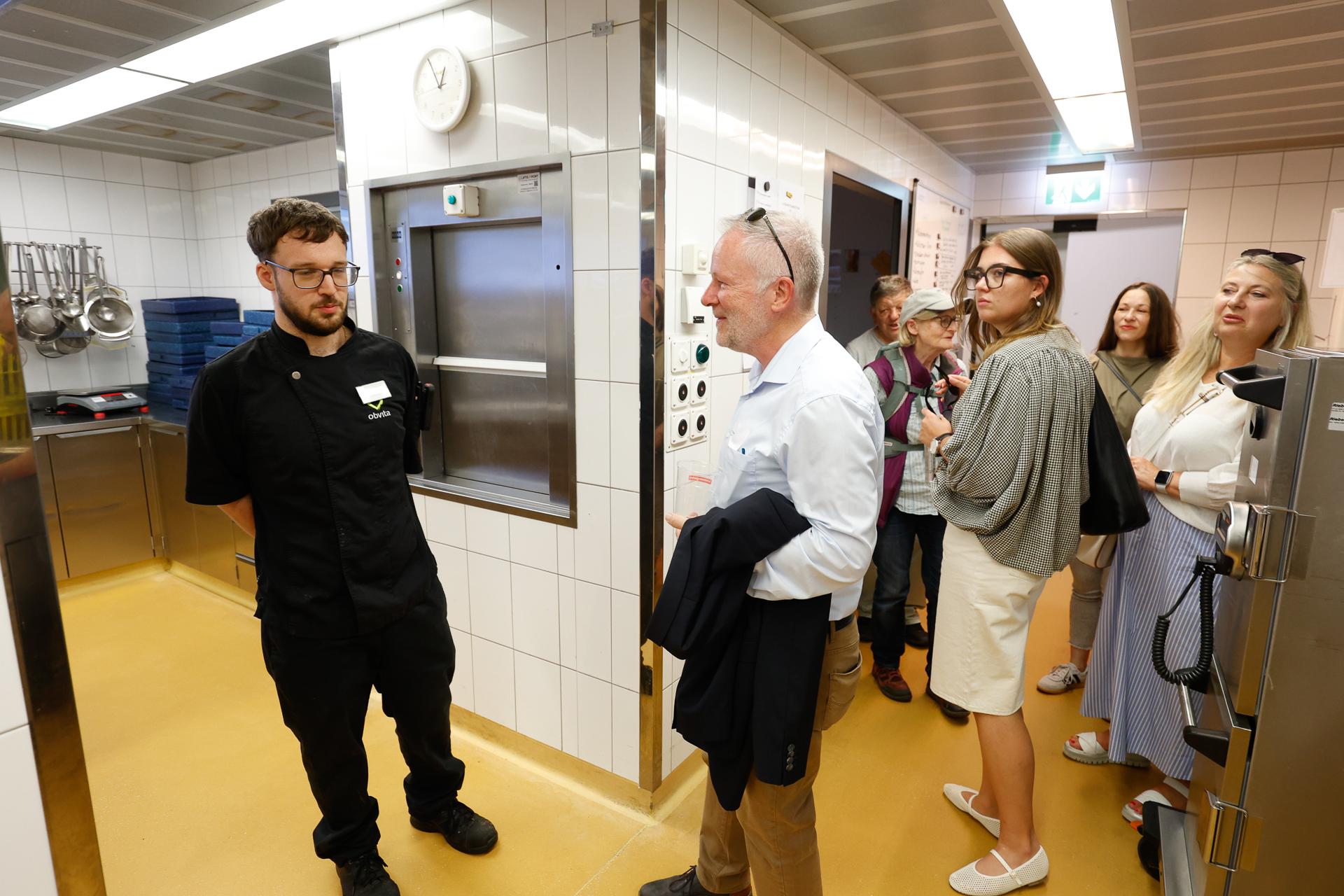 Eine Gruppe von Menschen hört einem Mann in schwarzer Kochkleidung zu, der in einer modernen, weiß gefliesten Küche spricht. An der Wand hängen Töpfe und Küchenutensilien, und über einem Ausgabeschalter ist eine Uhr zu sehen.