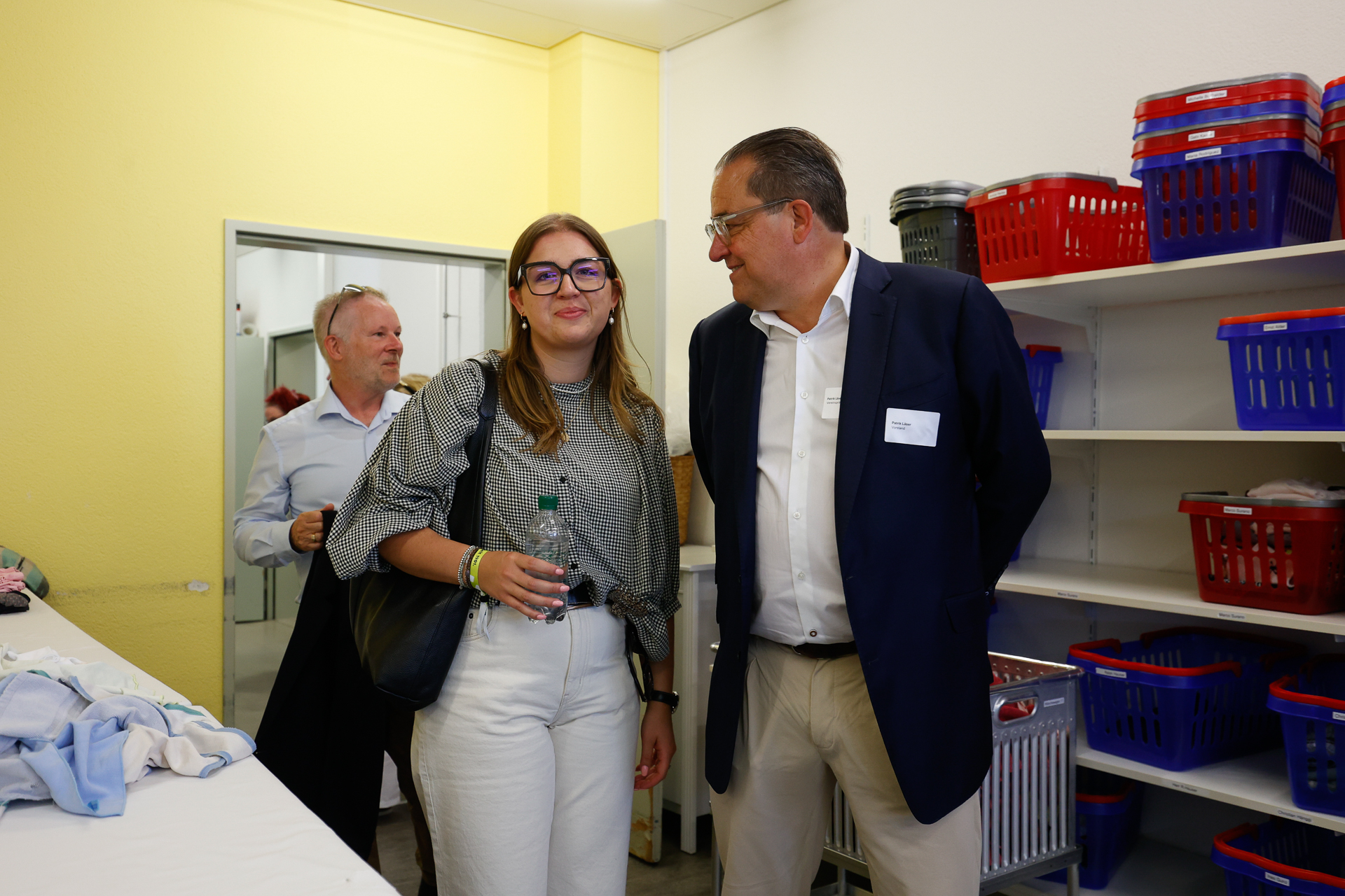 Drei Personen stehen in einem Waschraum, in dem Regale mit Wäschekörben stehen. Eine junge Frau mit Brille hält eine Wasserflasche in der Hand und lächelt, während zwei Männer in Anzügen daneben stehen; einer von ihnen hält eine Jacke in der Hand. Der Raum hat gelbe Wände und ist mit gefalteter Wäsche gefüllt.