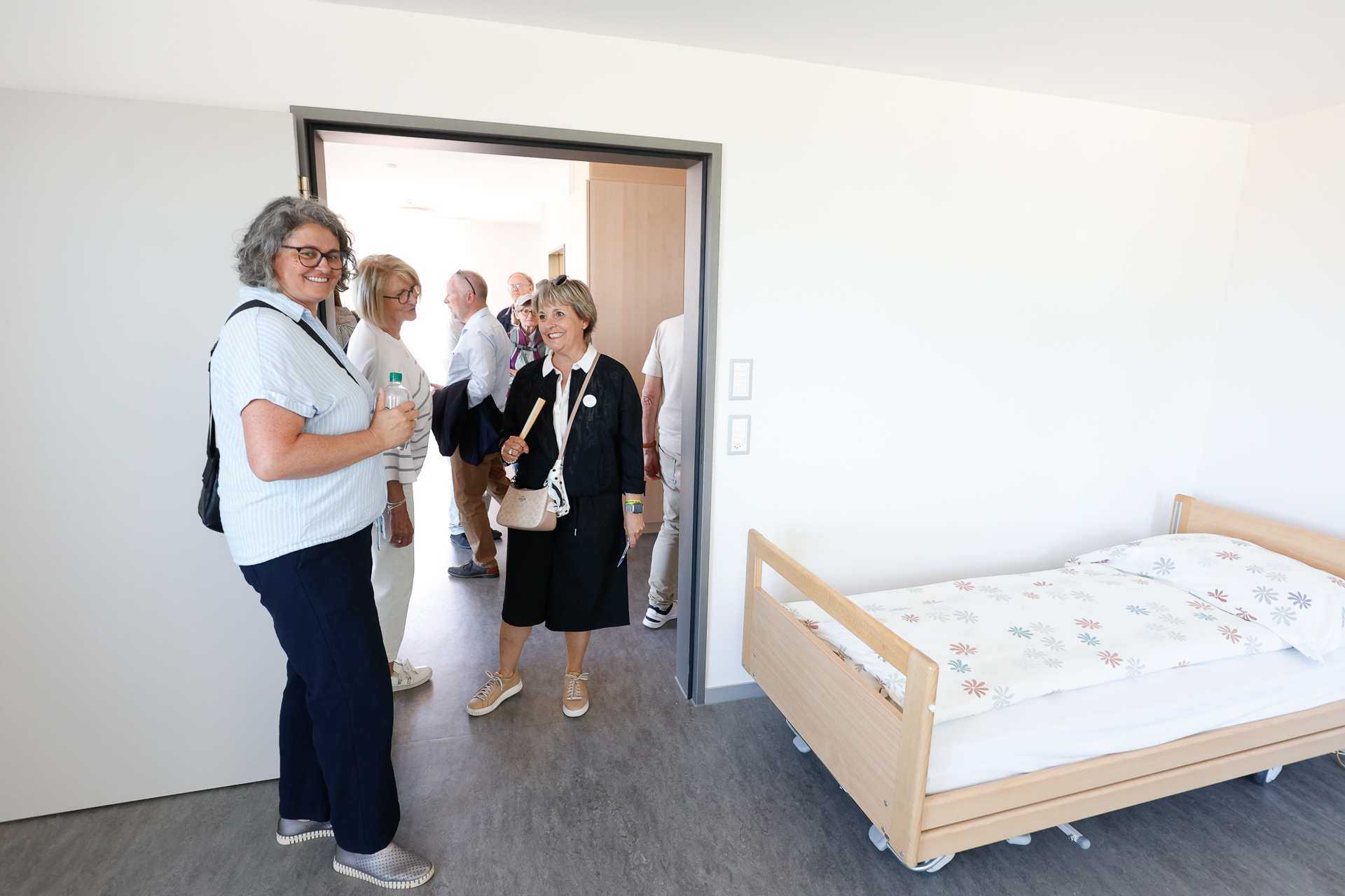 Eine Gruppe Erwachsener steht in der Tür und in einem hellen, aufgeräumten Zimmer mit einem Einzelbett. Die Atmosphäre wirkt freundlich, und einige der Anwesenden lächeln, während sie Taschen oder Wasserflaschen in den Händen halten.