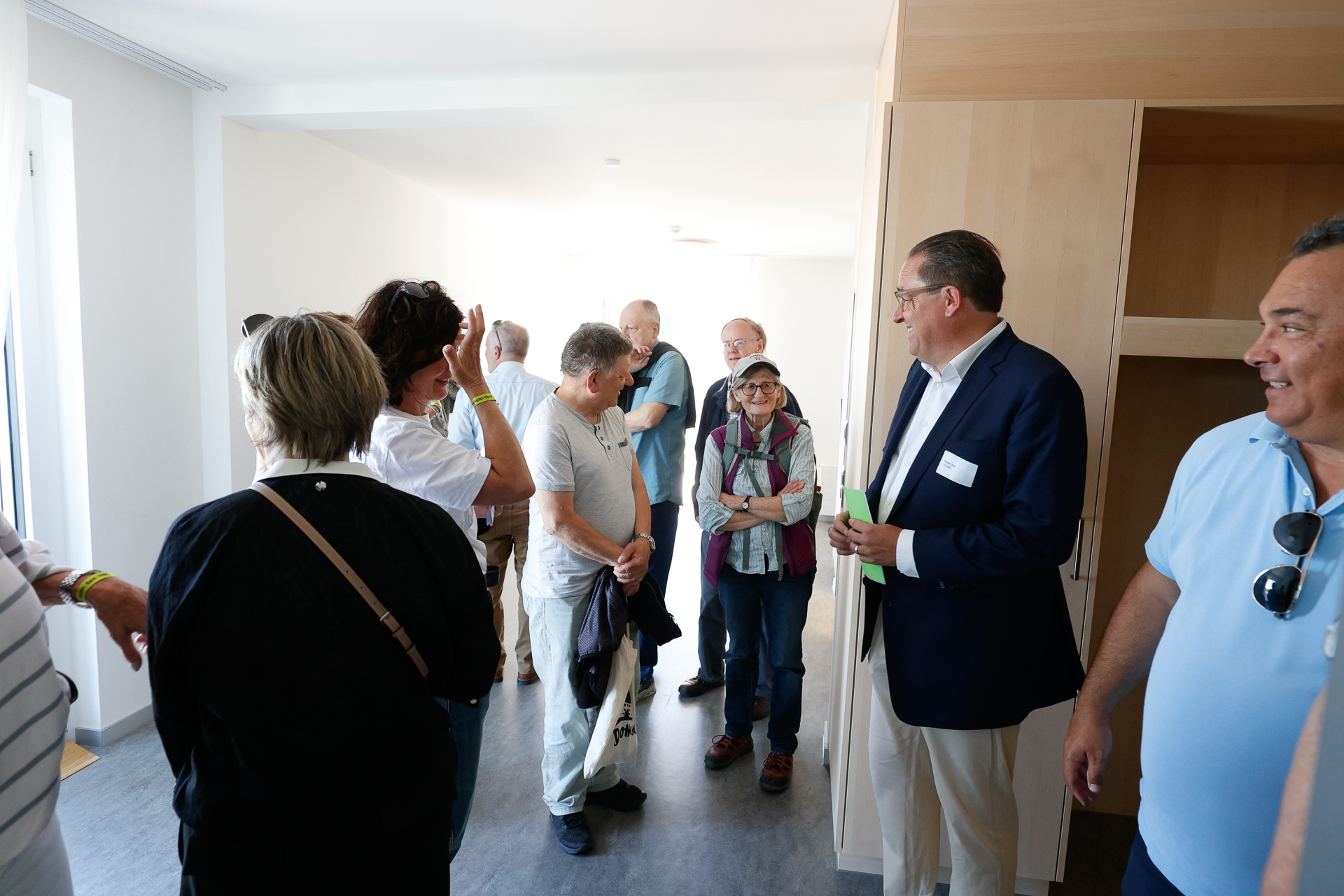 Eine Gruppe von Menschen, darunter Männer und Frauen, steht in einem hellen, unmöblierten Raum mit Holzregalen und unterhält sich. Ein Mann im Anzug scheint eine Diskussion oder eine Führung zu leiten.