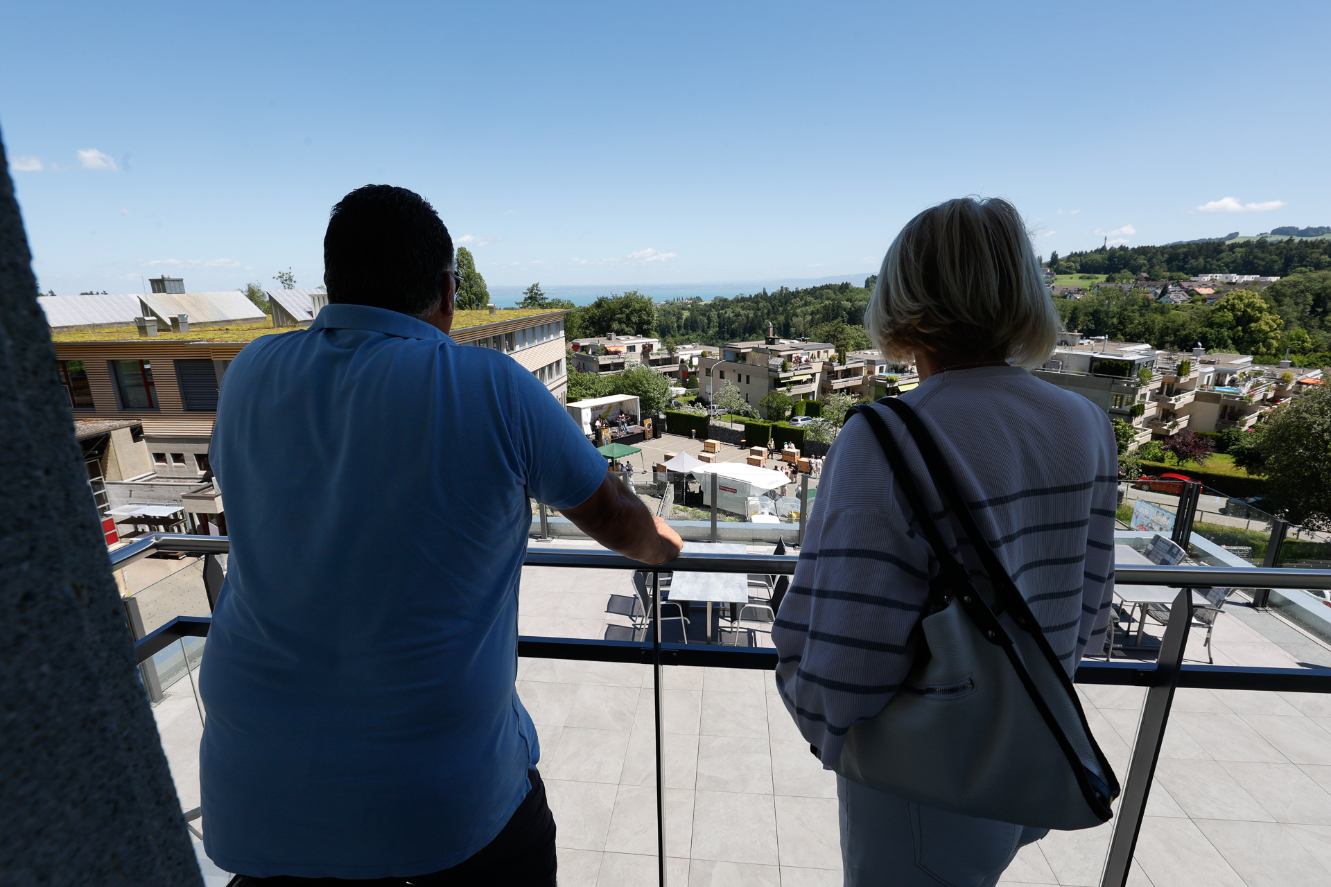 Zwei Personen stehen auf einem Balkon und blicken auf ein modernes Wohngebiet, im Hintergrund sind Grünflächen und ein strahlend blauer Himmel zu sehen. Sie stehen mit dem Rücken zur Kamera.