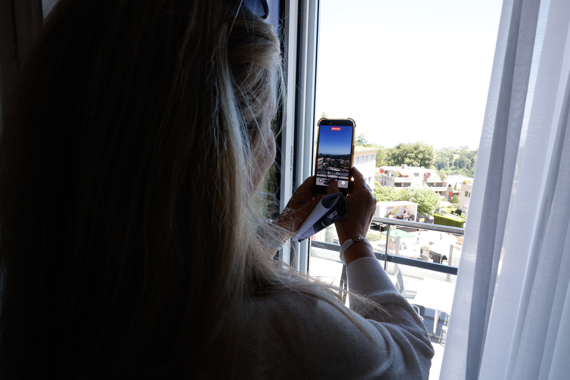 Eine Person mit langen, hellbraunen Haaren steht am Fenster und hält ein Handy in der Hand, um ein Foto von einer sonnigen Stadtkulisse im Freien mit Gebäuden und Grünflächen zu machen. Weiße Vorhänge sind teilweise beiseitegezogen.