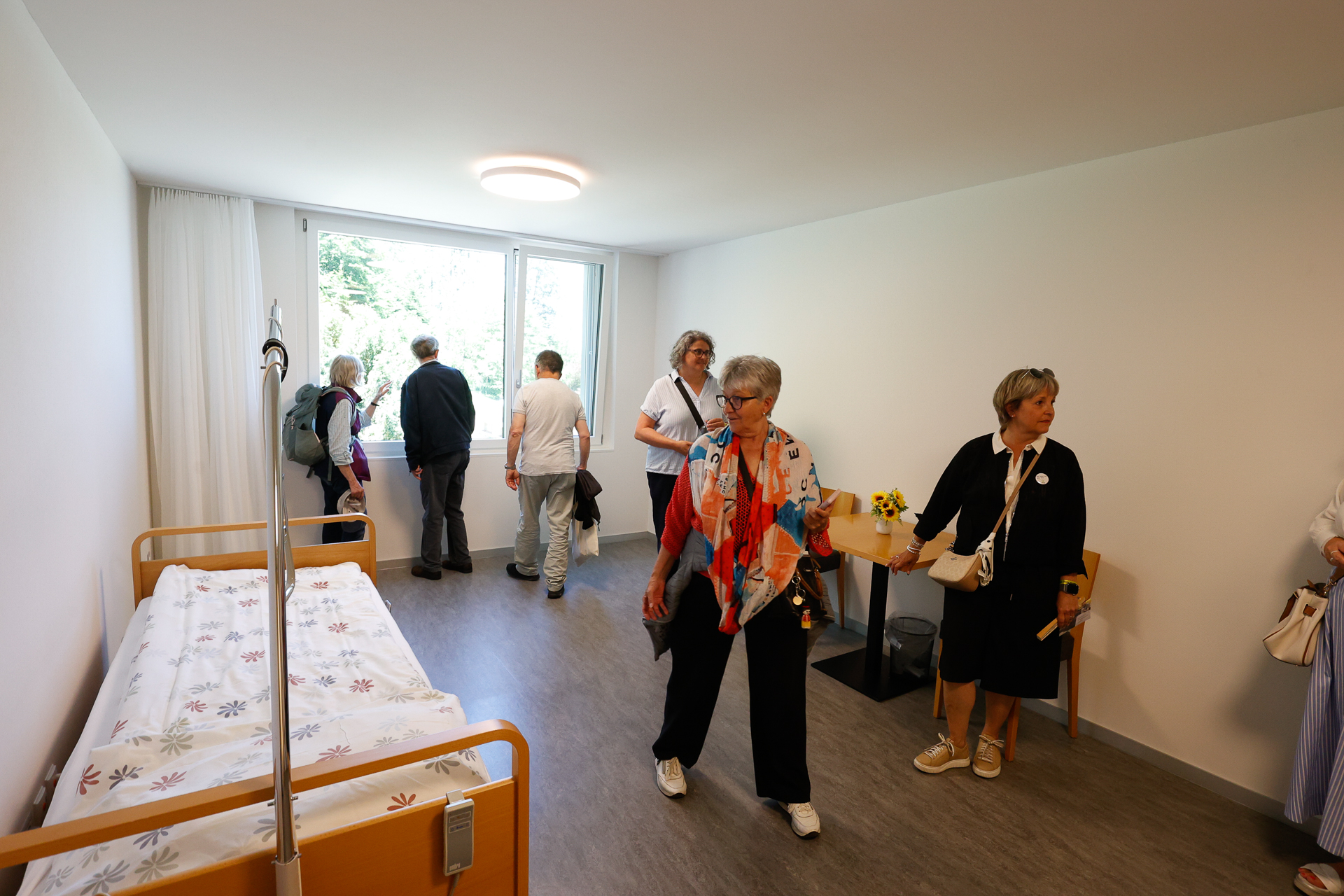 Eine Gruppe von Menschen, überwiegend ältere Erwachsene, steht und geht in einem schlichten, sauberen Raum umher, der mit einem Einzelbett, einem Schreibtisch, Stühlen und einem großen Fenster ausgestattet ist, durch das Tageslicht hereinfällt. Einige schauen aus dem Fenster, während andere sich unterhalten.