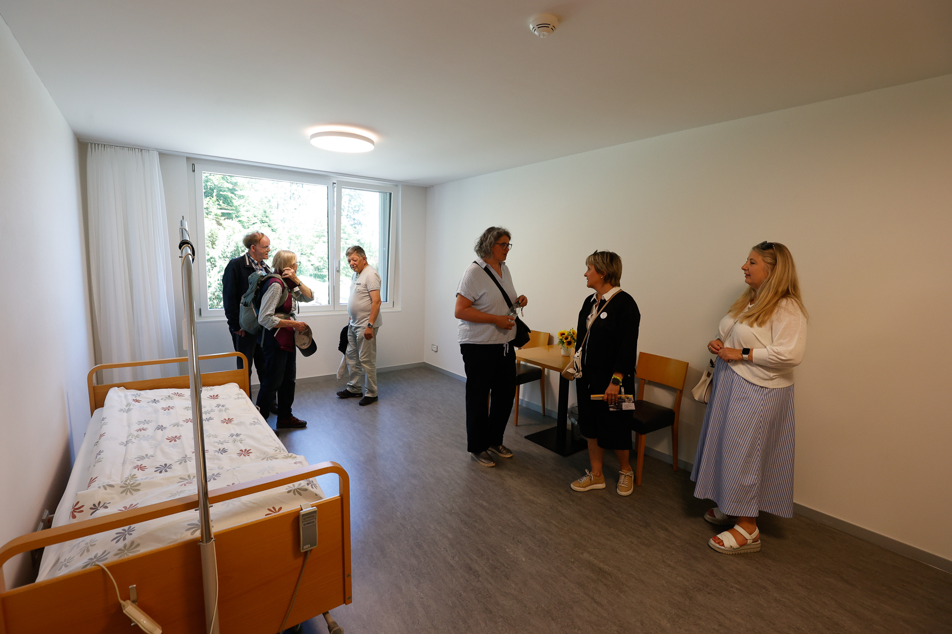 Sechs Menschen stehen in einem hellen, schlicht eingerichteten Krankenhaus- oder Pflegebereich und unterhalten sich. Dort stehen ein Einzelbett, ein kleiner Tisch mit Blumen und ein großes Fenster, durch das Tageslicht hereinströmt.